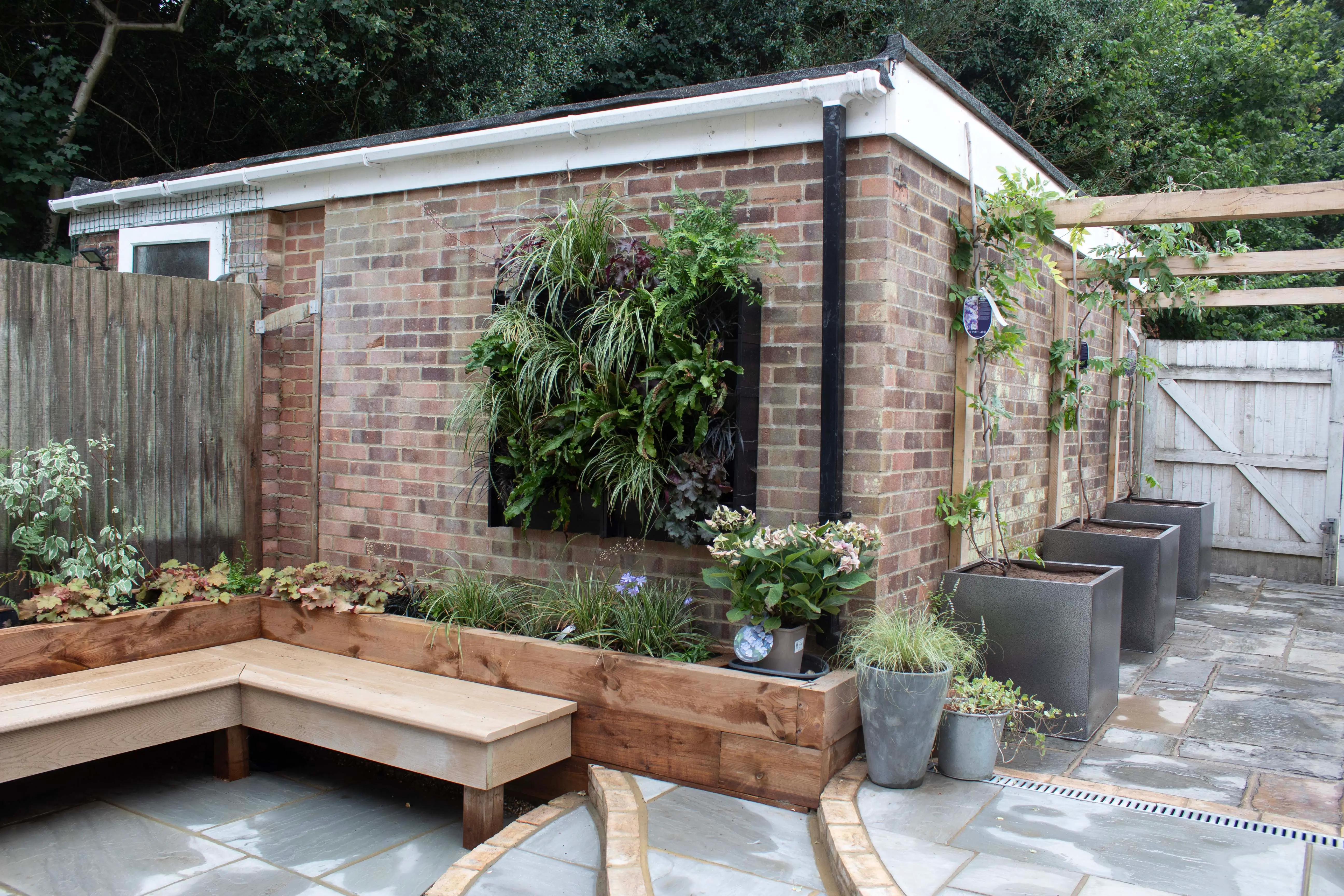 A cozy outdoor space features a wooden seating area and a brick structure with greenery and a trellis overhead.