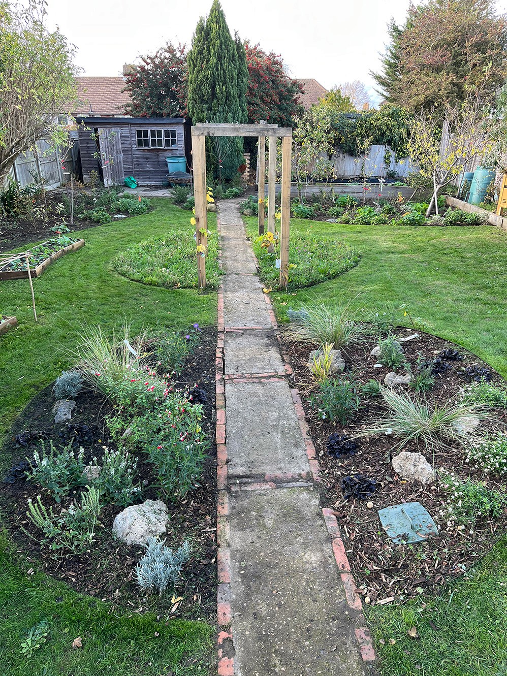 A stone pathway leads through a lush garden, framed by greenery and floral beds, toward an archway.