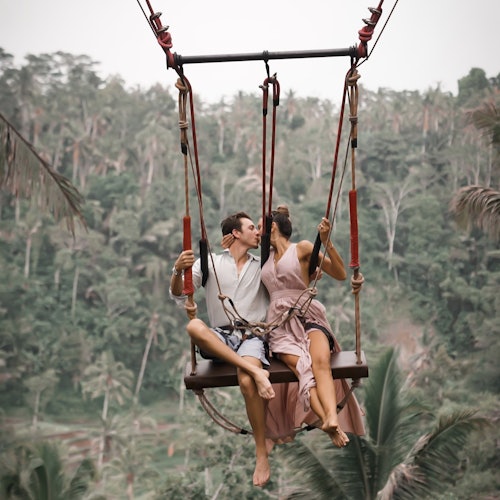 一對情侶坐在高高懸掛在茂密熱帶森林上的秋千上,互相親吻。