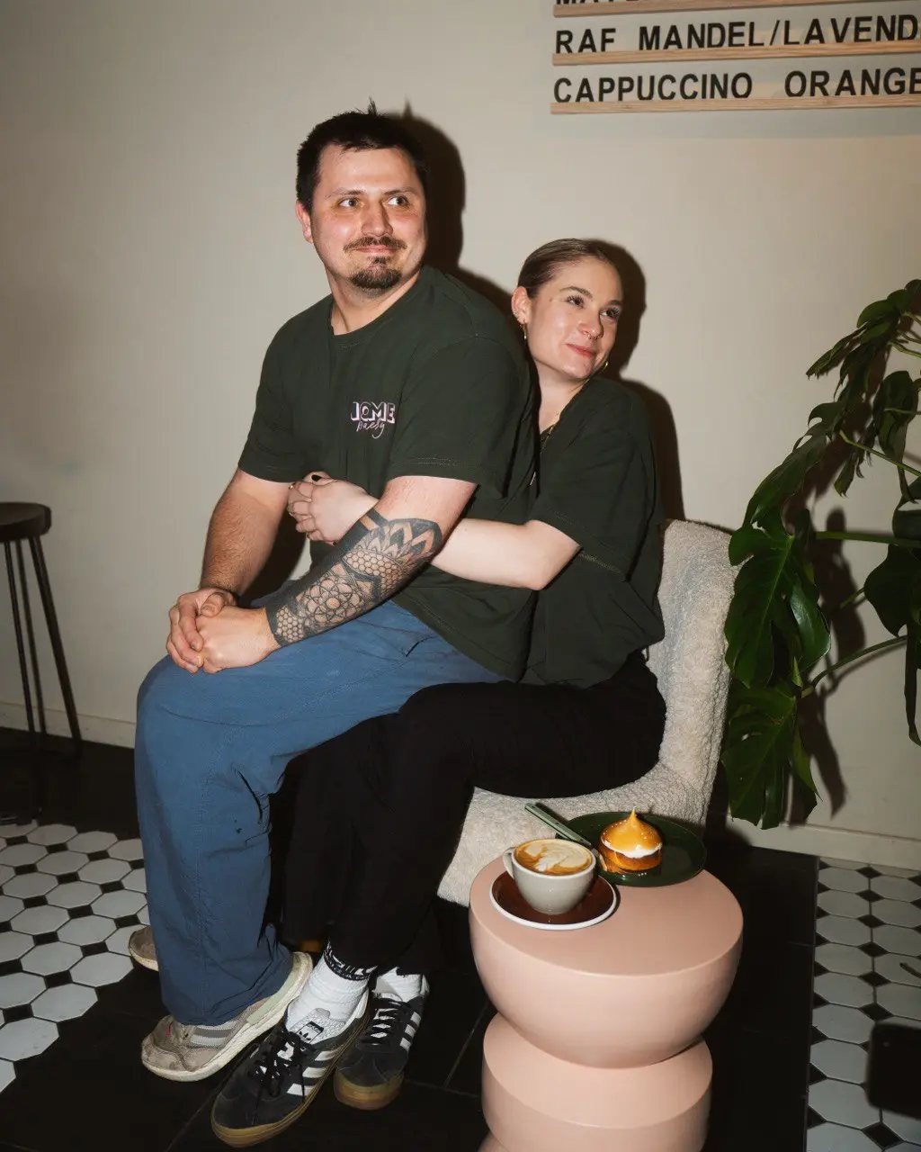 A person holds a cocktail glass containing a layered orange and yellow drink with a frothy top, set against a cozy bar background with warm lighting.