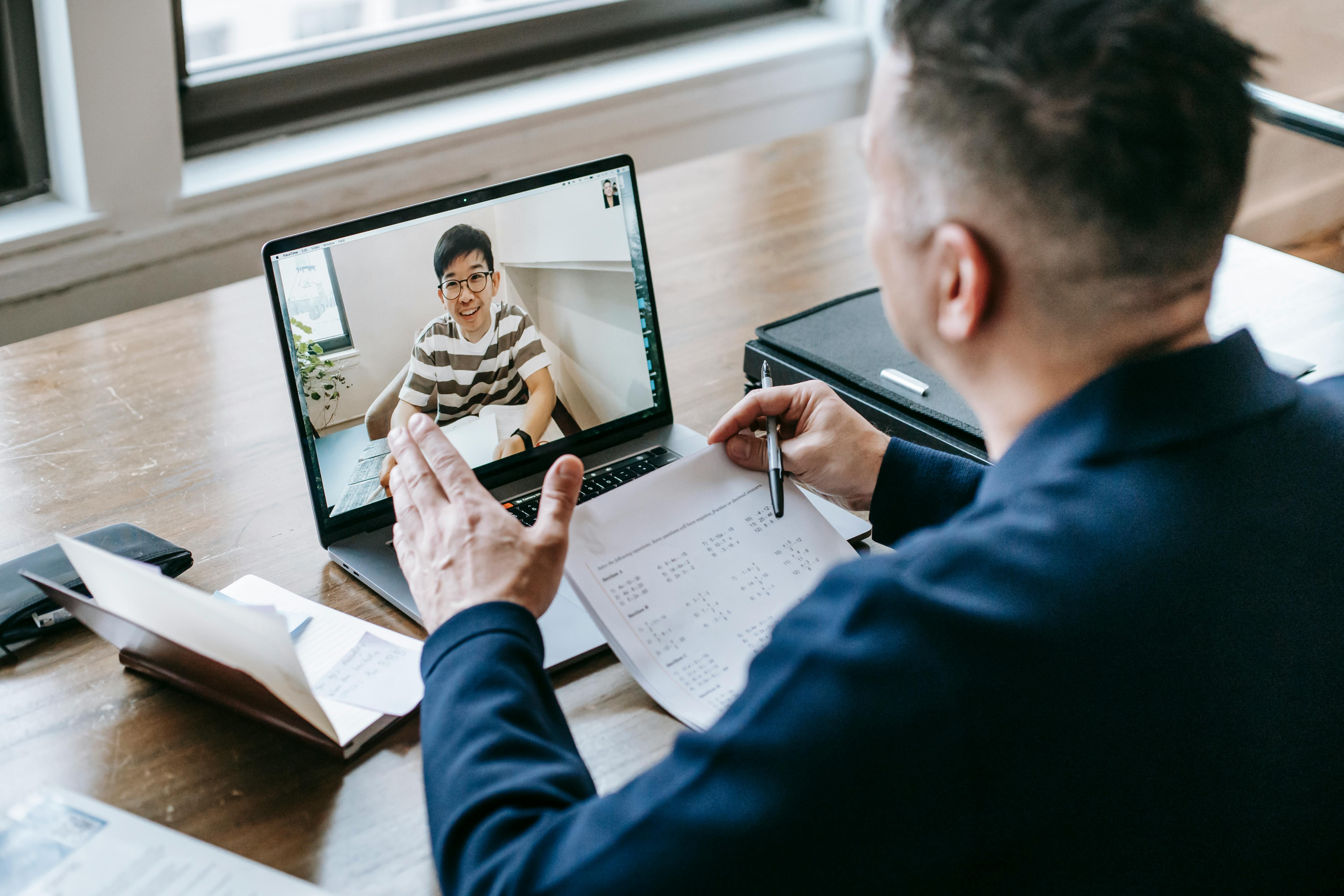  A man sitting at a desk with a laptop and a video call on the screen.