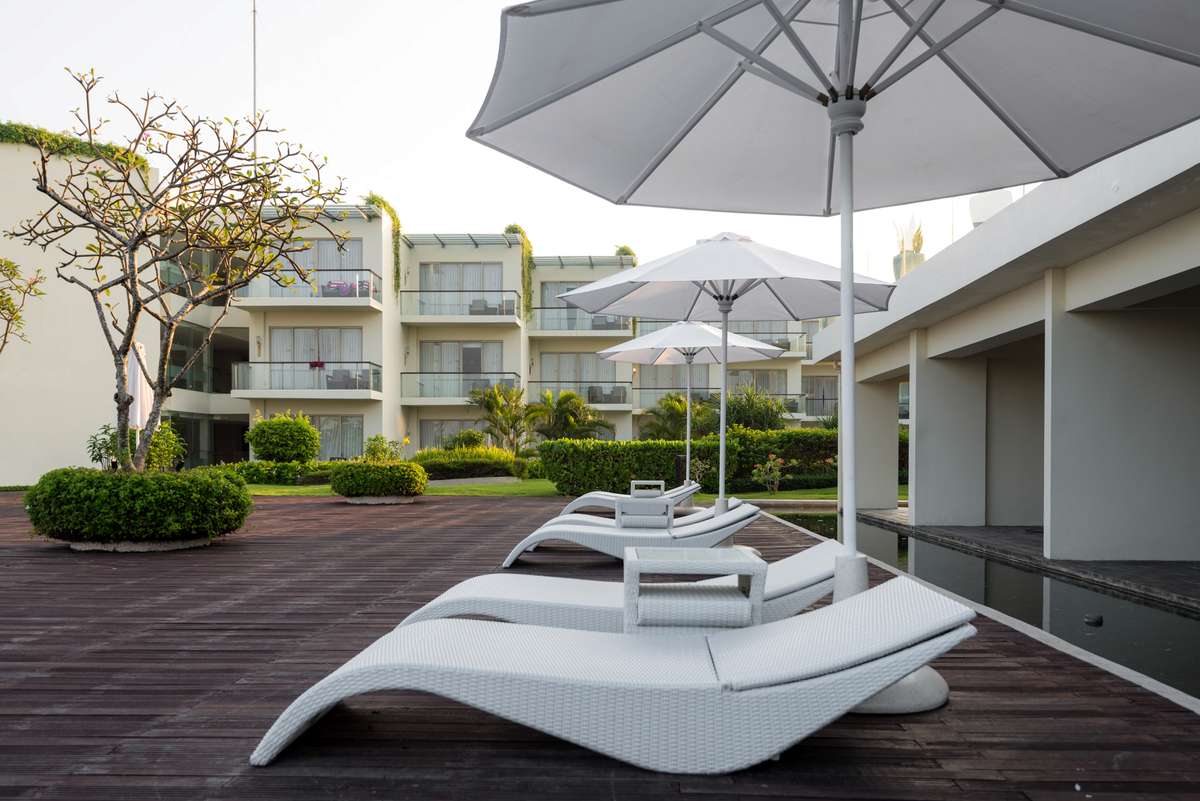 White lounge chairs and large patio umbrellas sit on a wooden deck in front of a modern resort building with balconies.