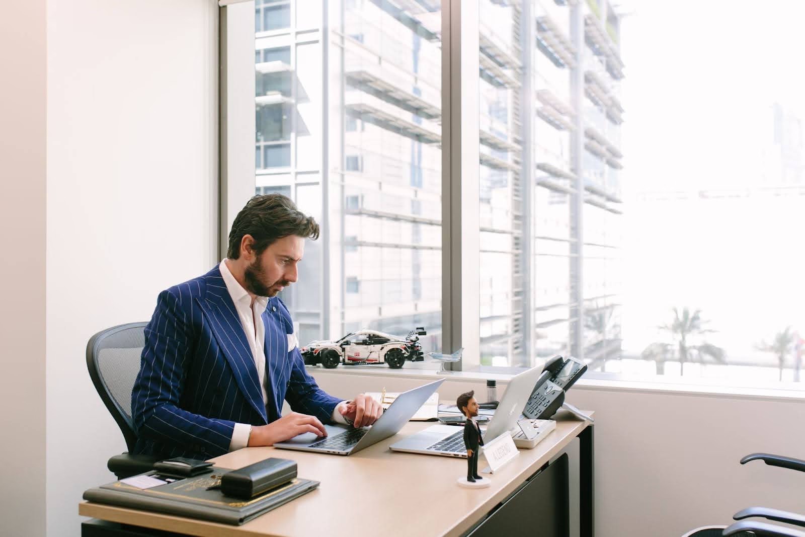 رجل أعمال ببدلة يعمل على جهاز لابتوب أمام نافذة مشمسة تطل على أفق المدينة، يجسد الكفاءة والنمو الاقتصادي المرتبط بخطوات تأسيس شركة في دبي