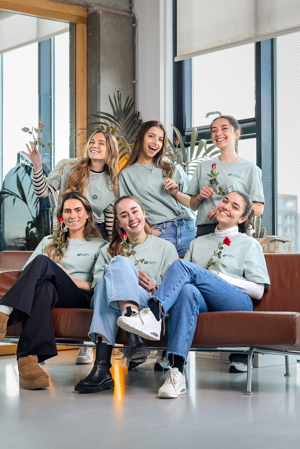 A group of six women, casually dressed, holding red roses in a bright room