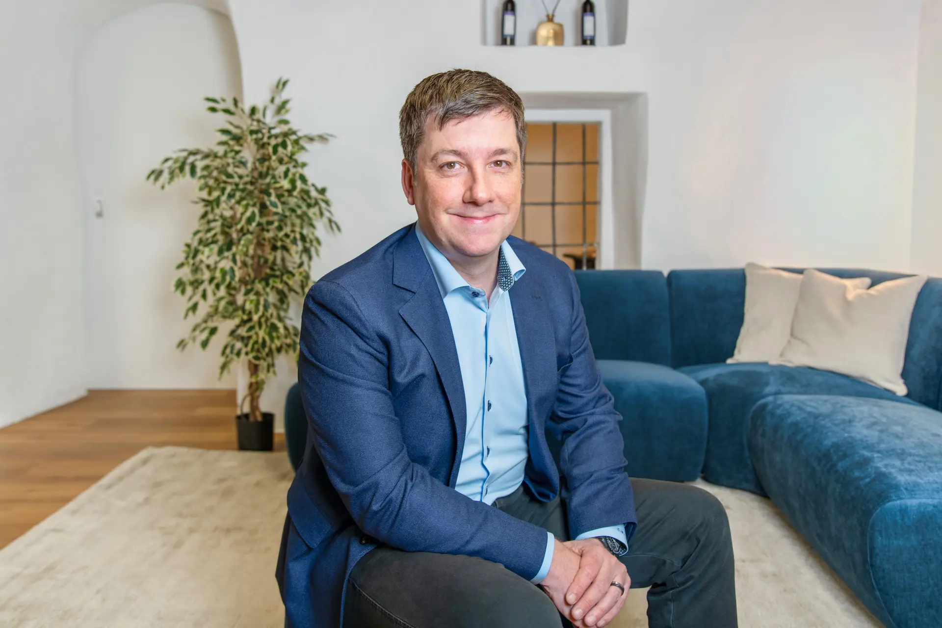 Immobilienmakler von Kocher Immobilien AG in einem blauen Anzug sitzt lächelnd vor dem blauen Sofa in der Immo Lounge