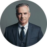 An old man wearing a formal suit and tie, standing confidently with a neutral expression.