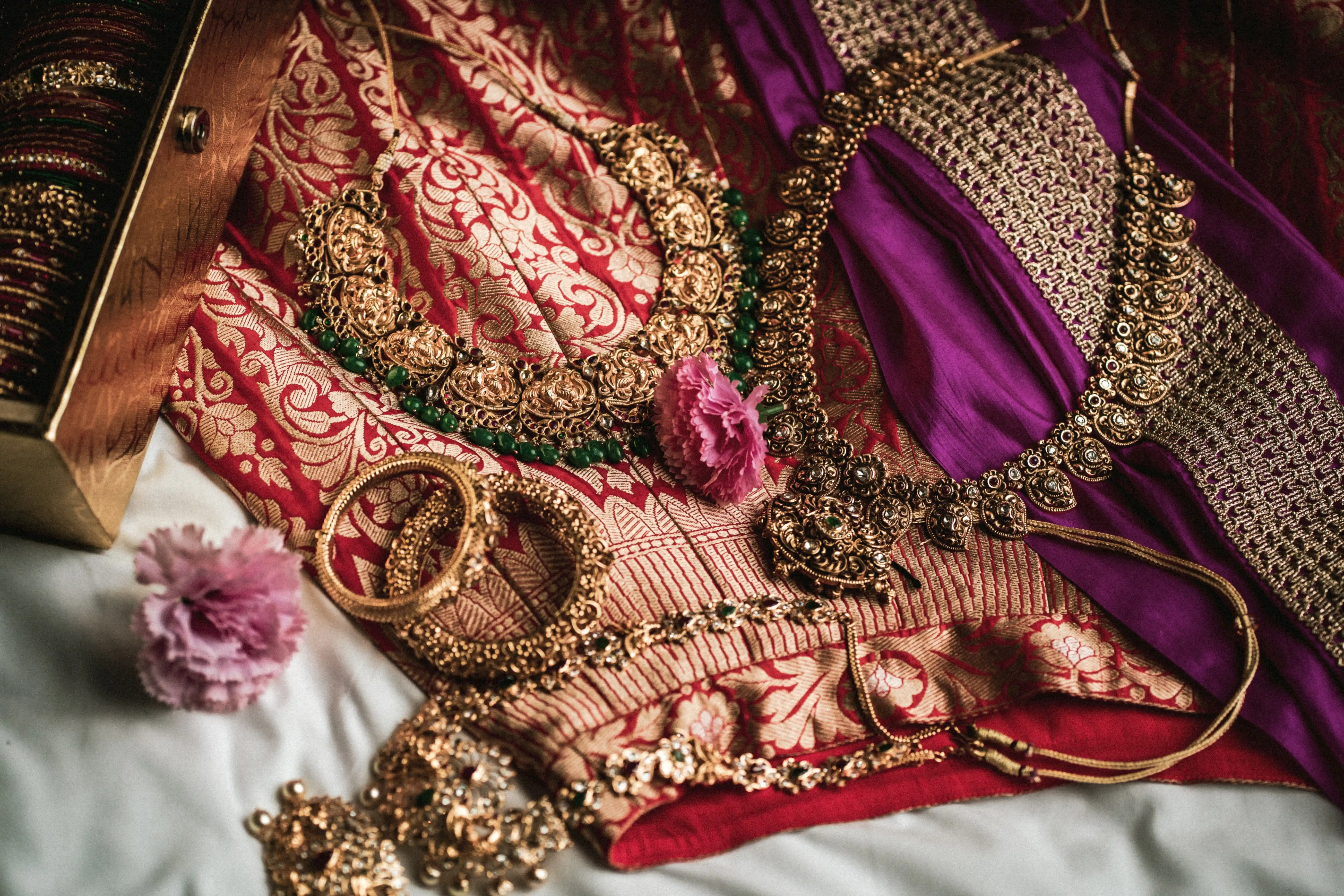 Fine art close-up of traditional Indian bridal jewelry, including gold necklaces and bangles, arranged on a richly patterned red saree, beautifully styled by Out of The Blues Fine Art Wedding Photography in Hyderabad.