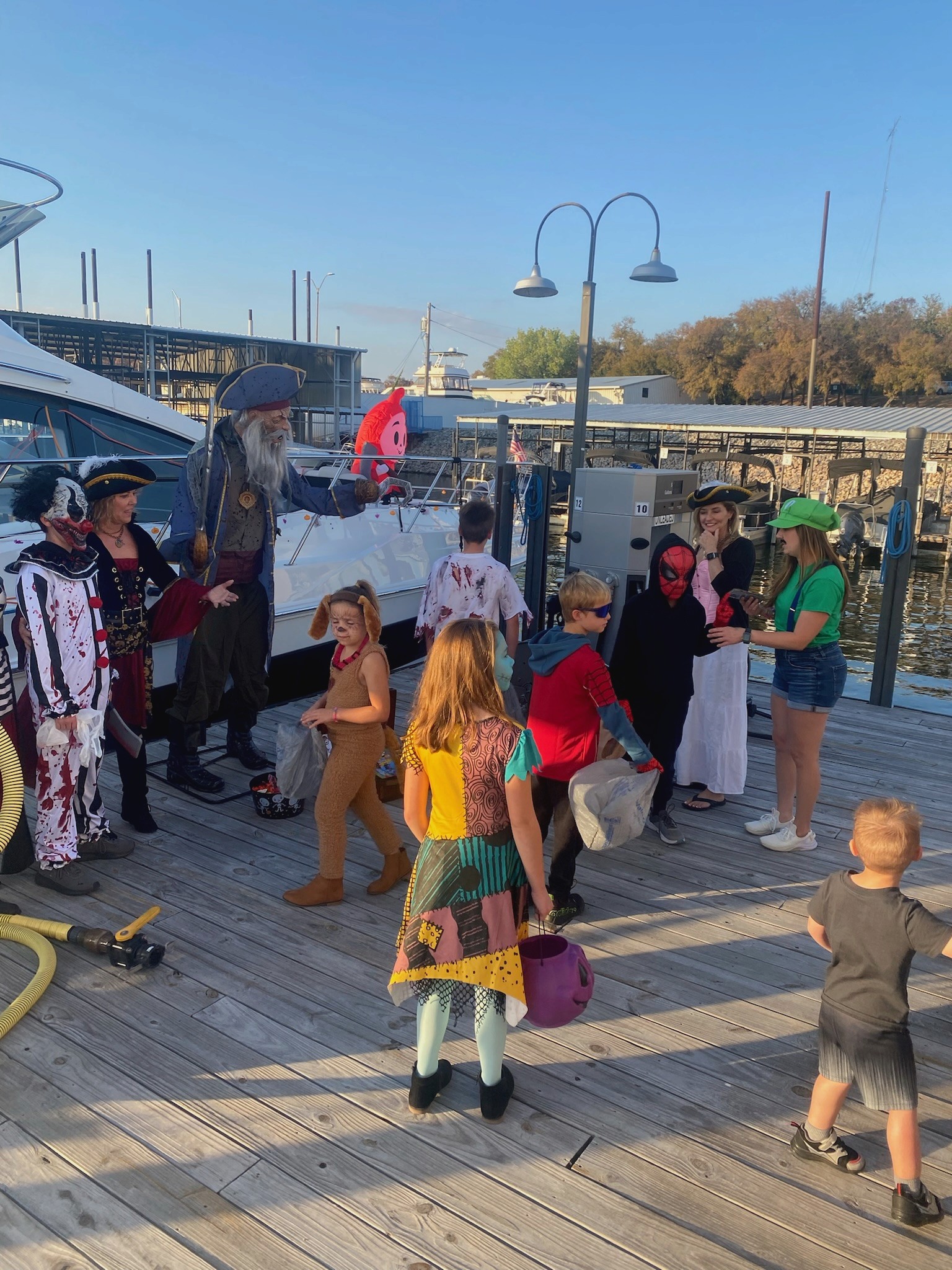 A diverse group of children and adults, dressed in various colorful costumes, gather joyfully on a wooden dock by the marina under clear blue skies, suggesting a festive or themed event.