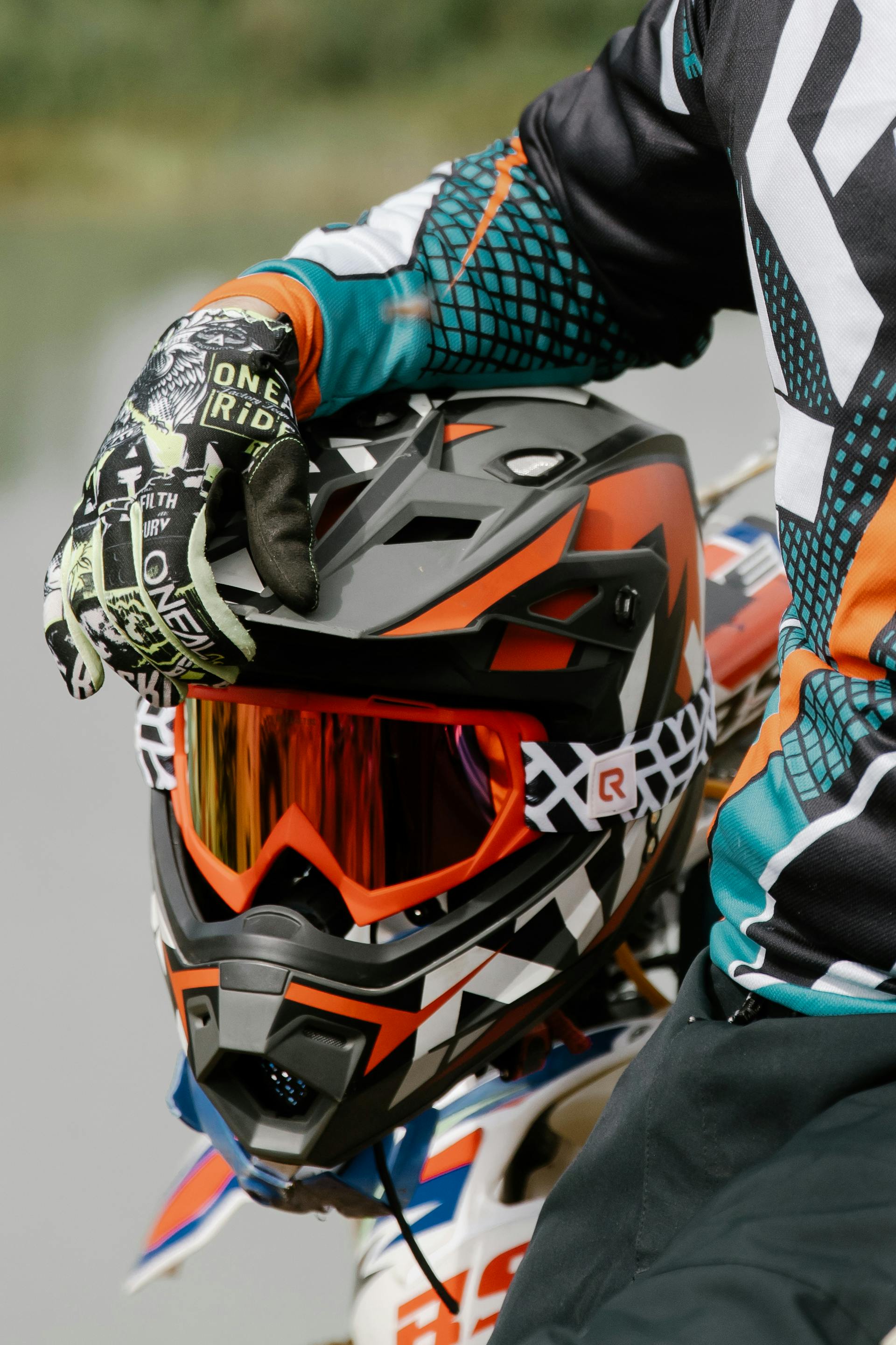 A detailed shot of a black and orange motocross helmet with red-tinted goggles, with a rider’s gloved hand resting on the visor.