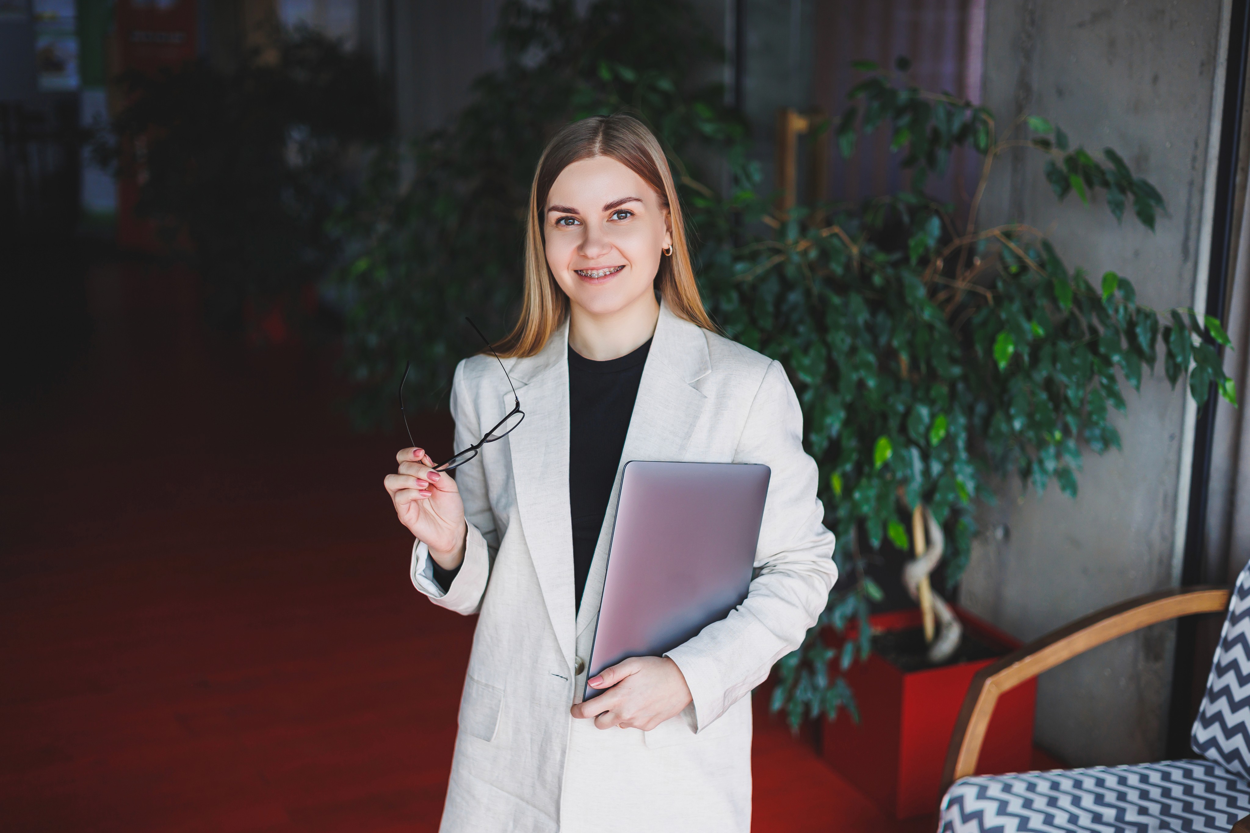 Young woman with long hair, wearing a white blazer, holds glasses and a laptop, smiling confidently. Red carpet and greenery in the background.
