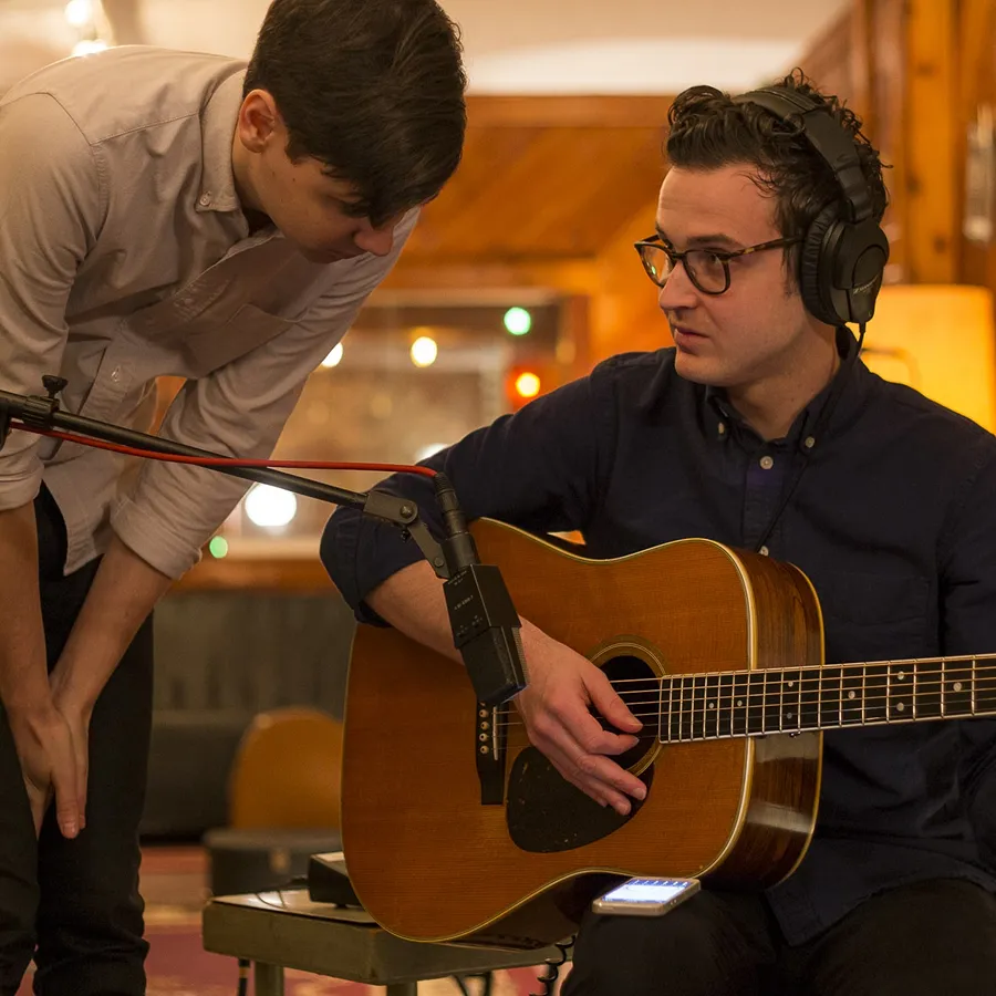 Two people in a warm, cozy setting; one is tuning a guitar while the other observes intently.