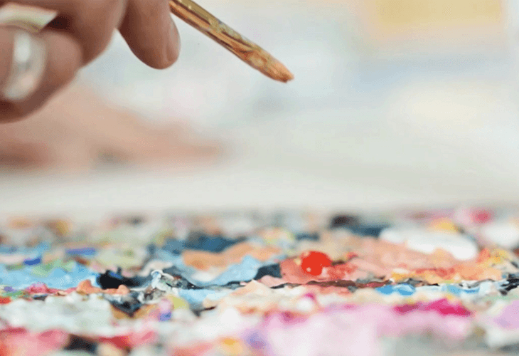 A hand holds a wooden-handled paintbrush, applying paint to a colorful, textured surface that appears to be a painting or artwork. The surface is covered with small, multicolored paint swatches and has a slightly uneven, layered appearance. The background is blurred and light-colored.