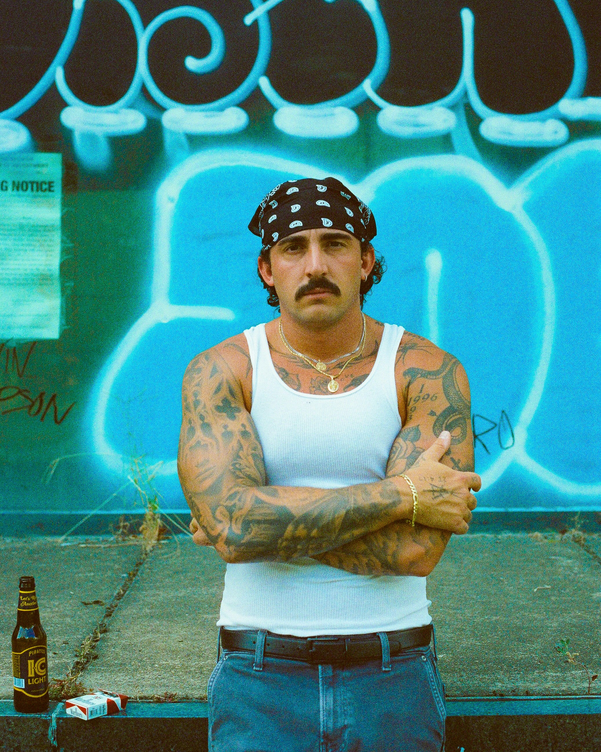 Man with bandana and tattoos stands against graffiti wall.