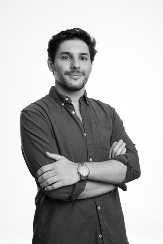 Black-and-white studio portrait of a smiling man with short dark wavy hair and light stubble, wearing a dark button-up shirt with an open collar and a wristwatch, arms crossed, against a plain light background.