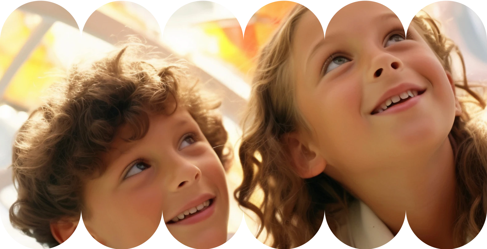 Close-up de duas crianças, um menino e uma menina, com cabelos cacheados/ondulados, olhando para cima com sorrisos, em um ambiente bem iluminado.