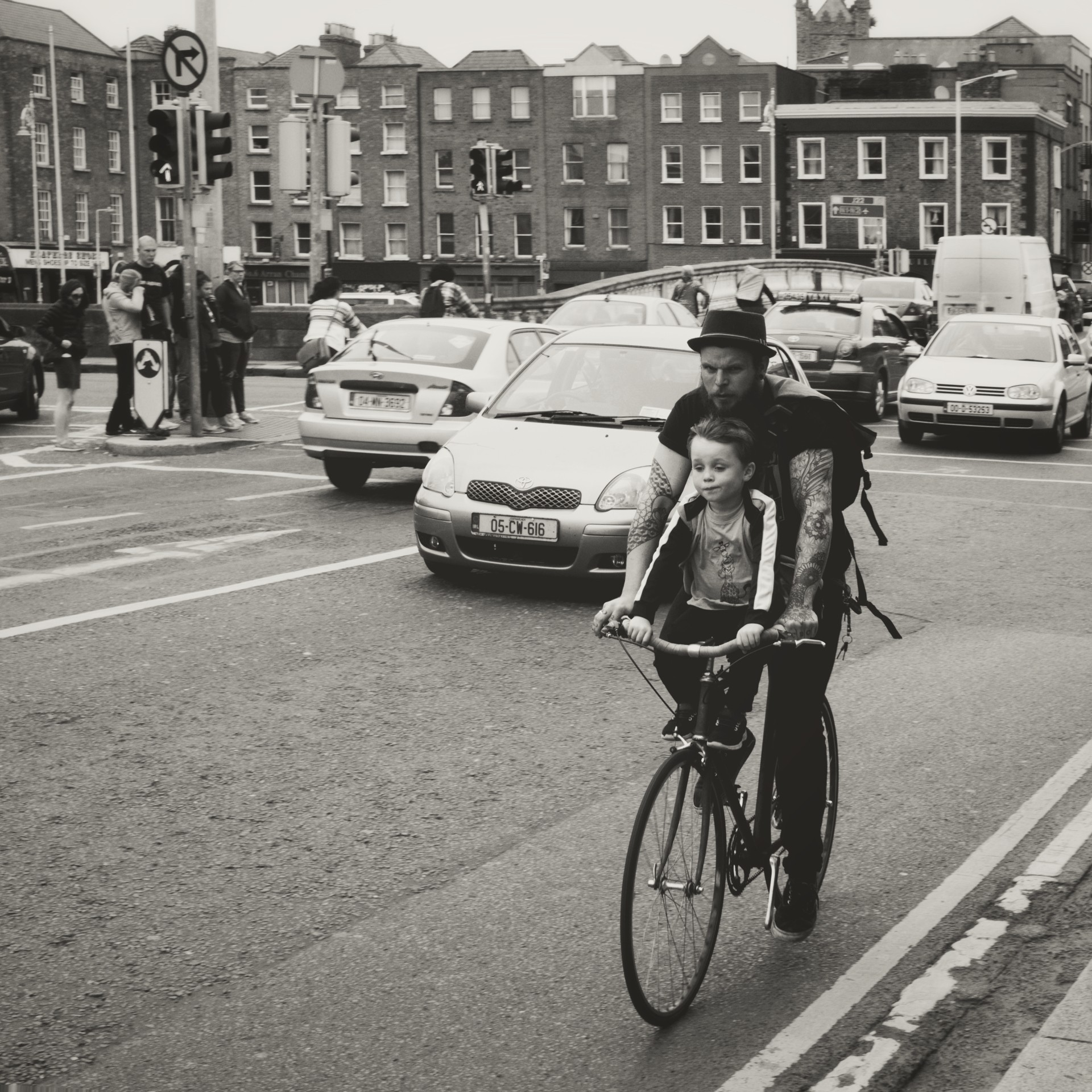 Urban black-and-white scene showing everyday city life: a parent and child sharing a cargo bike on a sidewalk next to a brick wall, framed by traffic and pedestrians.