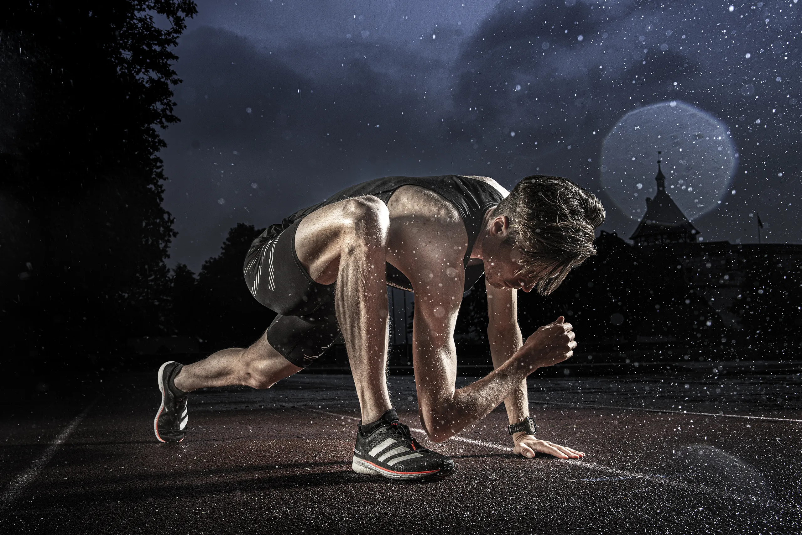 A powerful shot of a male sprinter in the starting blocks on a wet track at night with water droplets caught in the light.