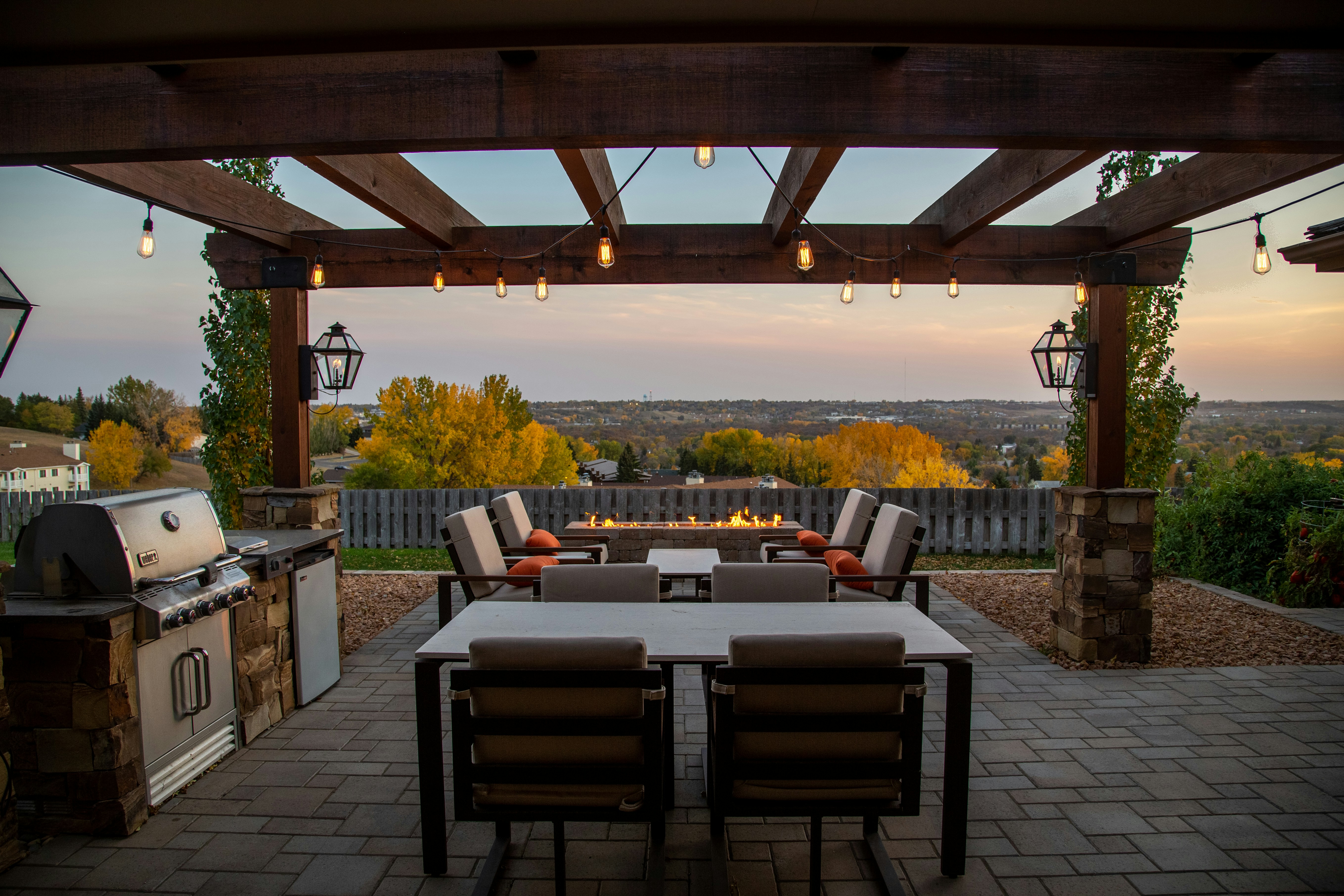 This outdoor patio features a cozy dining and seating area under a wooden pergola with string lights, offering a picturesque view of a scenic landscape.