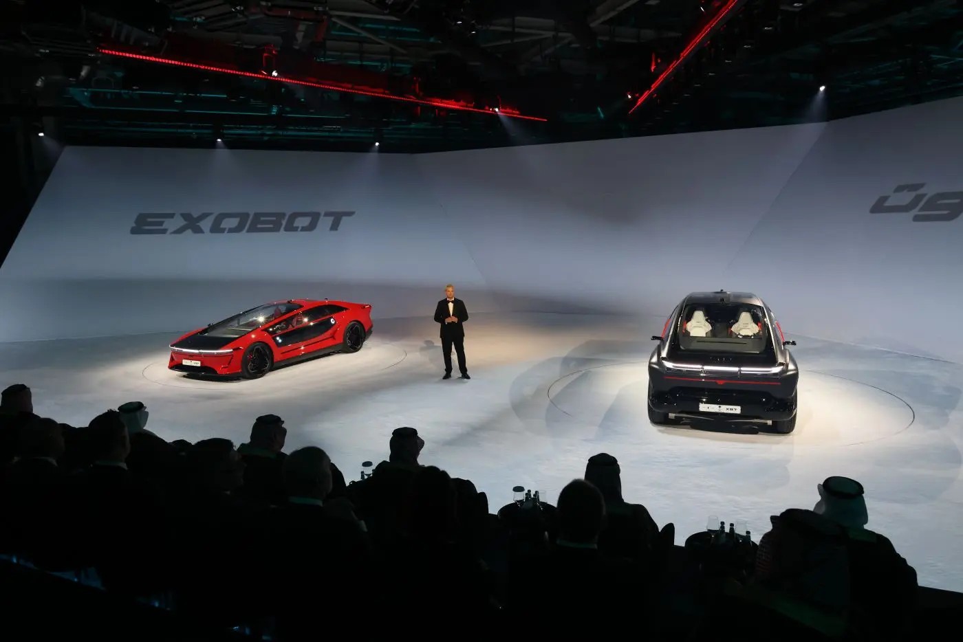 A red EXOBOT sedan and a dark SUV on a lit stage as a speaker presents to a seated audience