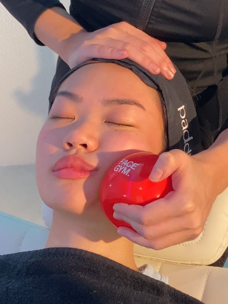 A person receiving a facial treatment at Face Gym, with a trainer using a red facial tool on their cheek, promoting facial fitness.