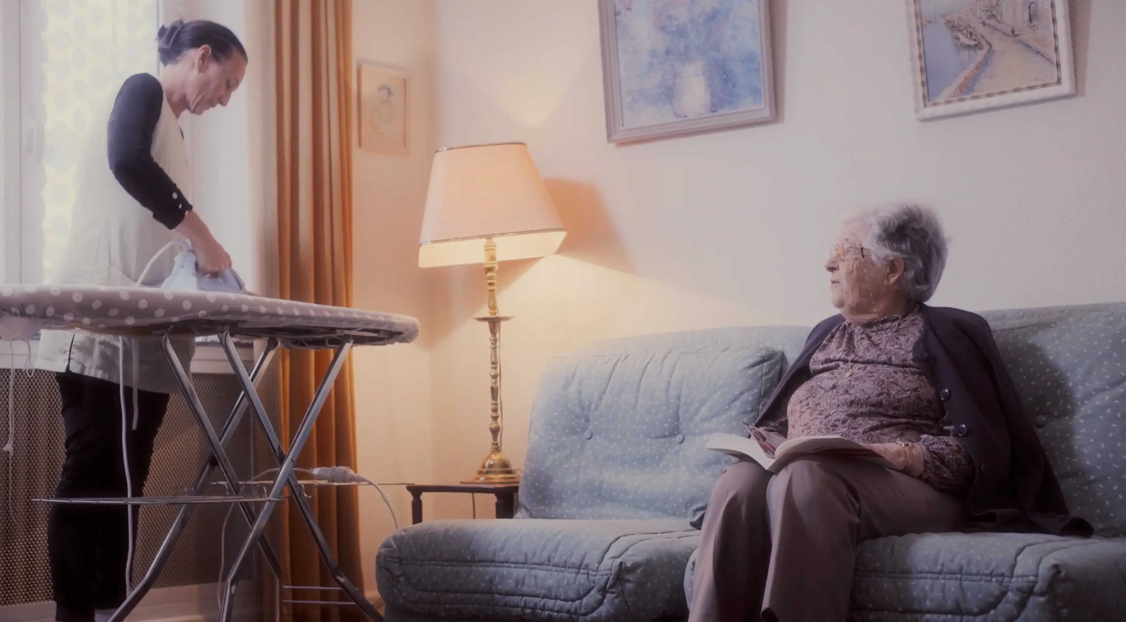 An elderly person sits on a sofa reading a book, while another person stands nearby ironing clothes next to a warm-lit lamp in a cozy living room adorned with framed artwork on the walls.