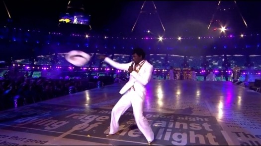 A performer in a white suit energetically dances on a brightly lit stage, tossing a white hat amidst a colorful, illuminated backdrop during a lively concert.