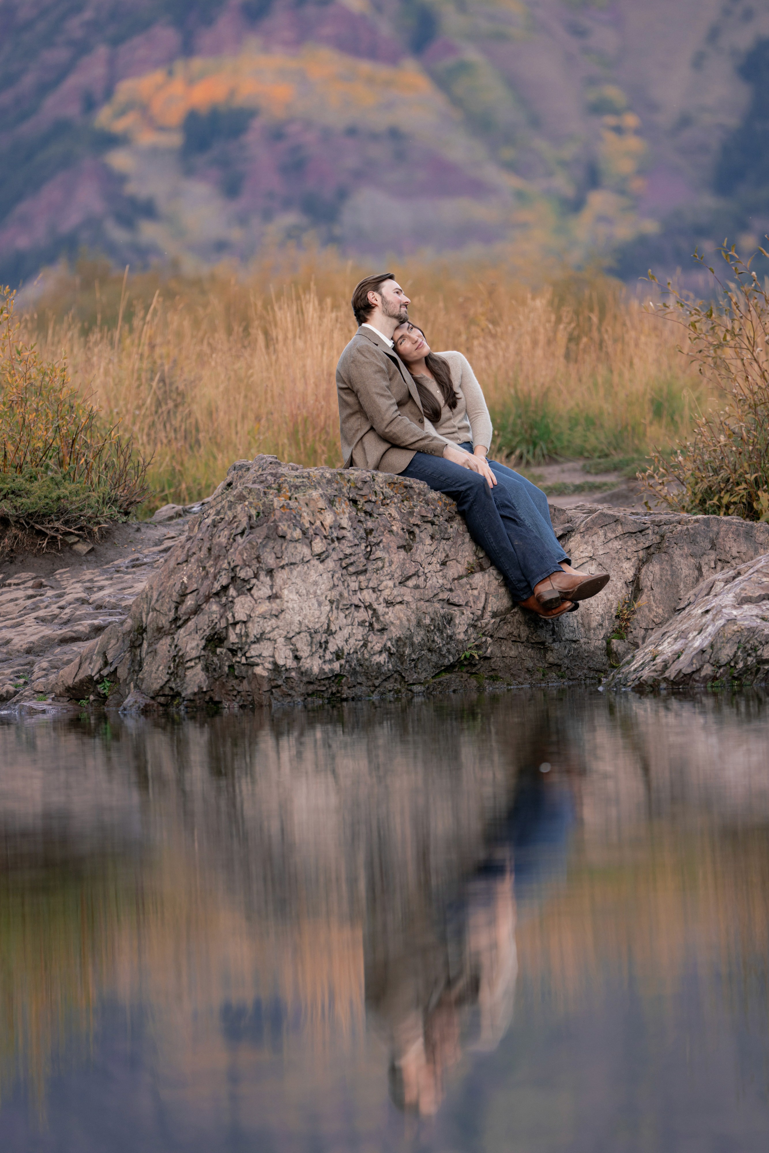 Lexi rests her head on Tim’s shoulder as they sit beside still water reflecting their portrait.