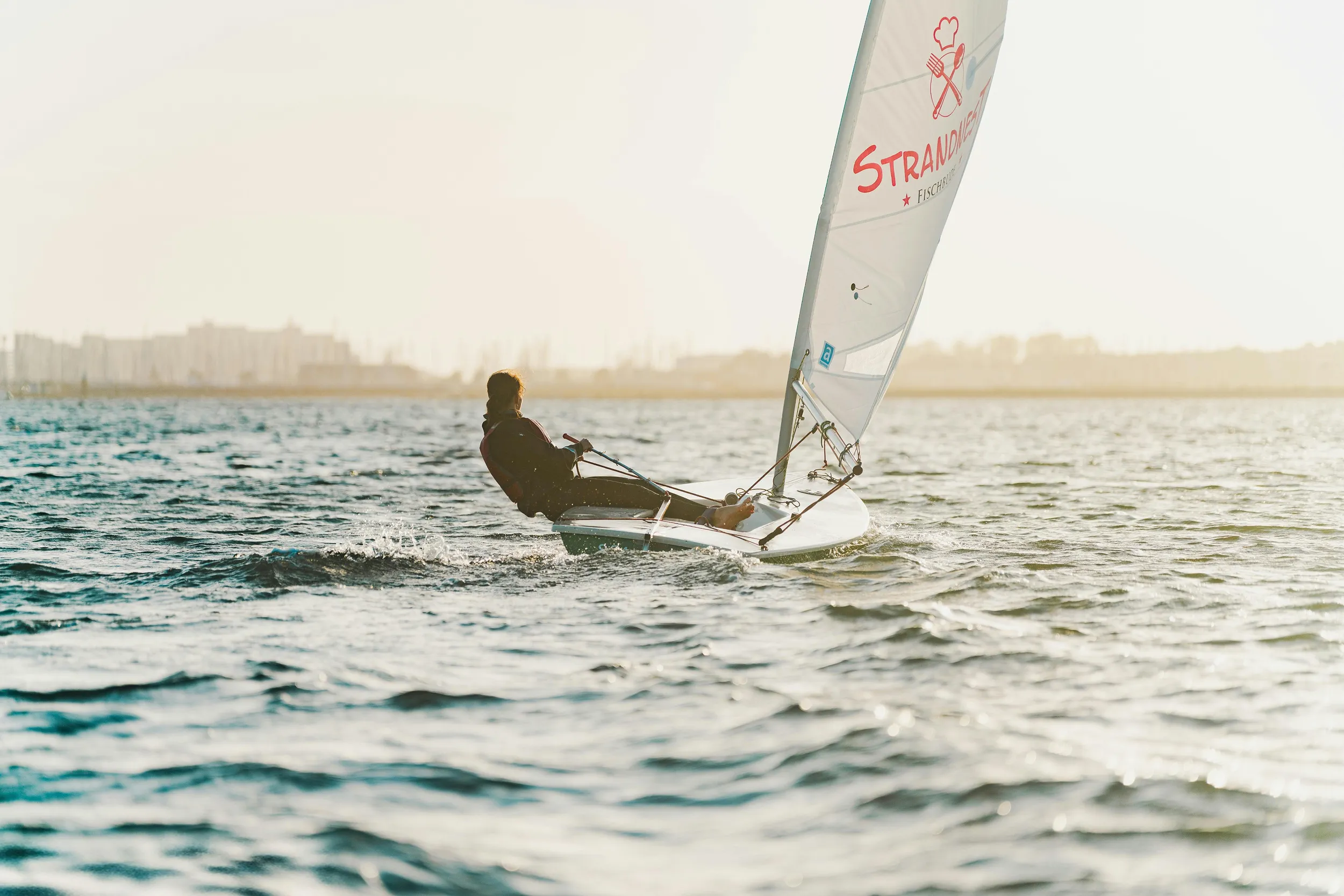 Solo sailor navigating a small sailboat on open water at sunset, outdoor adventure lifestyle