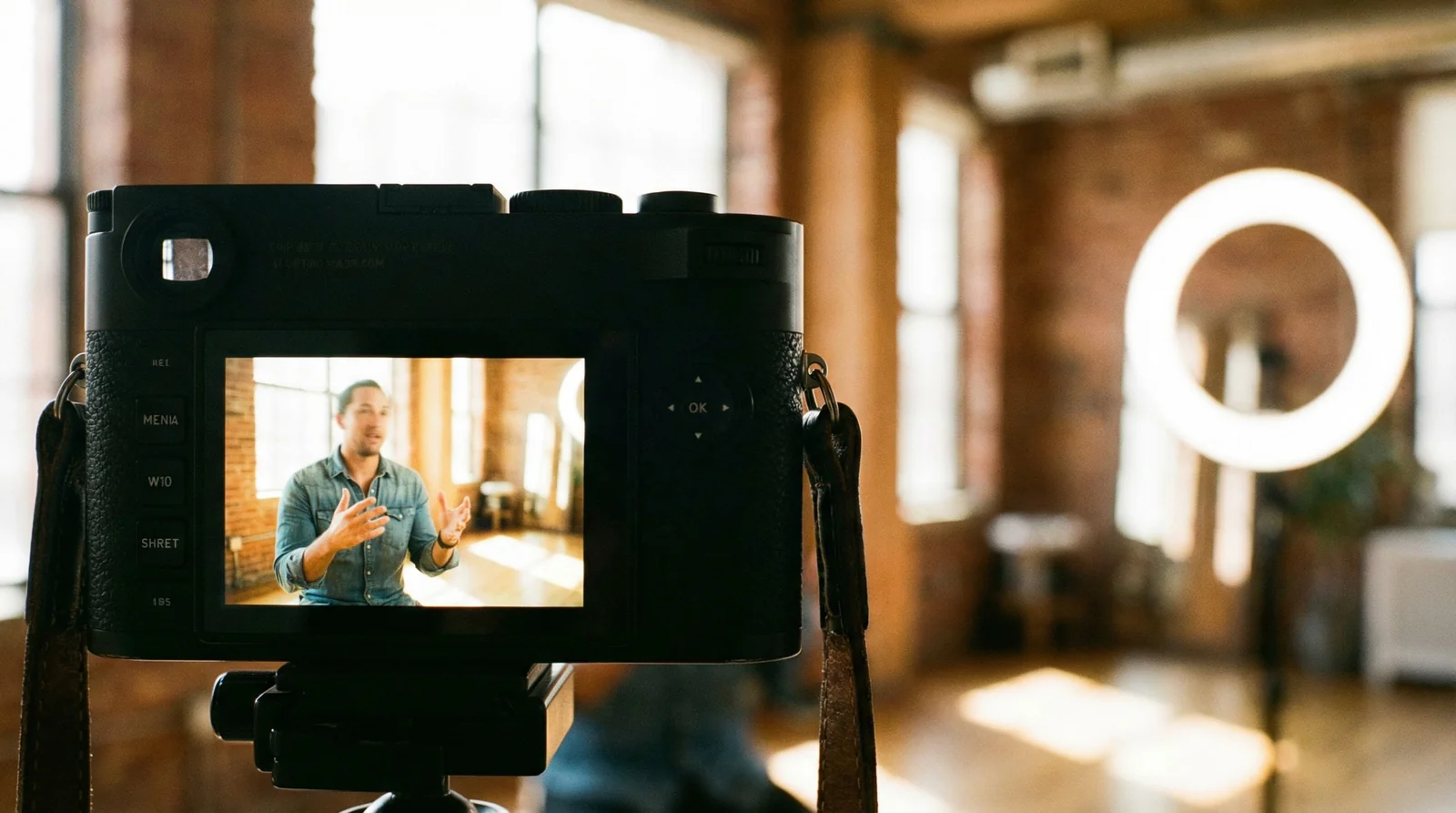 Camera viewfinder showing recording in progress, camera body sharp in foreground, ring light and loft with exposed brick softly in background