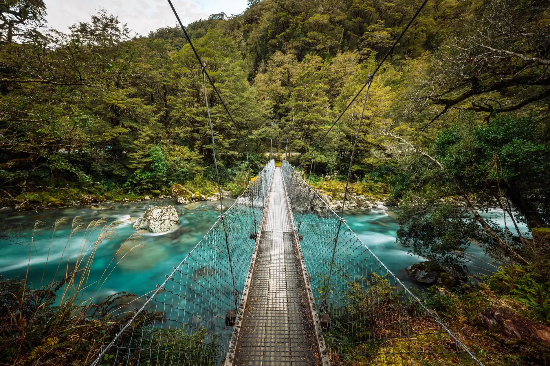 A suspension footbridge crossing a bright turquoise river in a forested gorge
