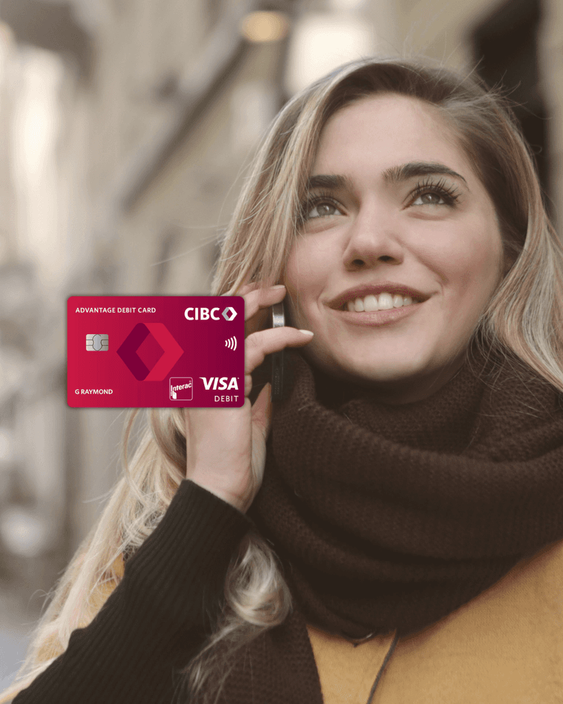 A smiling young woman outdoors in winter clothing talking on her smartphone, with a red CIBC Advantage Visa Debit card overlaid, representing the CIBC Welcome to Canada Banking Package and big-bank reliability for new immigrants.