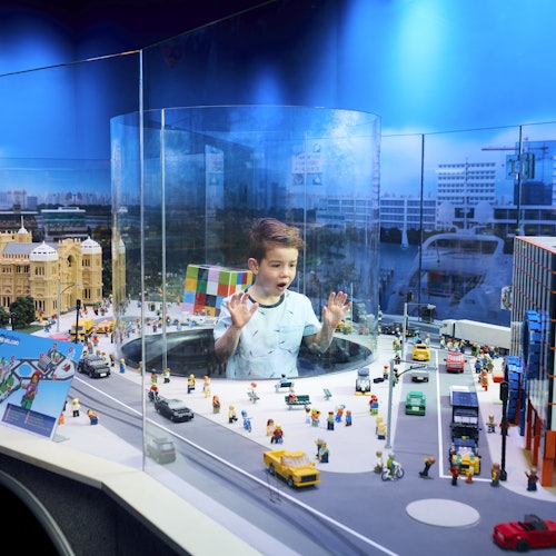 Dječak se nalazi unutar prozirnog cilindra u izlošku Lego grada, okružen zgradama, vozilima i Lego figuricama.