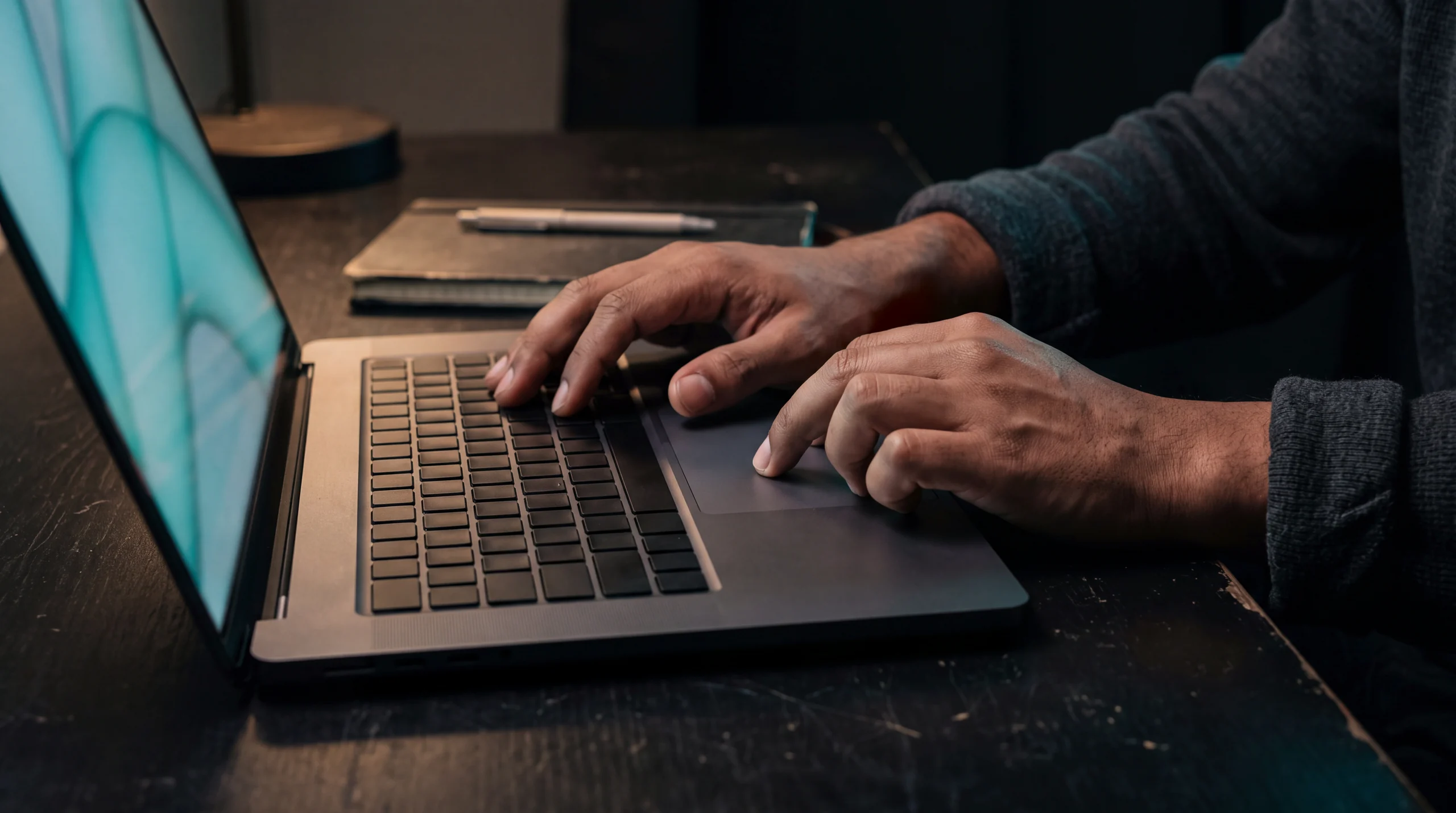 Mains posées sur le clavier et le pavé tactile d'un ordinateur portable dont l'écran affiche des formes turquoise abstraites : l'humain pilote l'outil d'intelligence artificielle.
