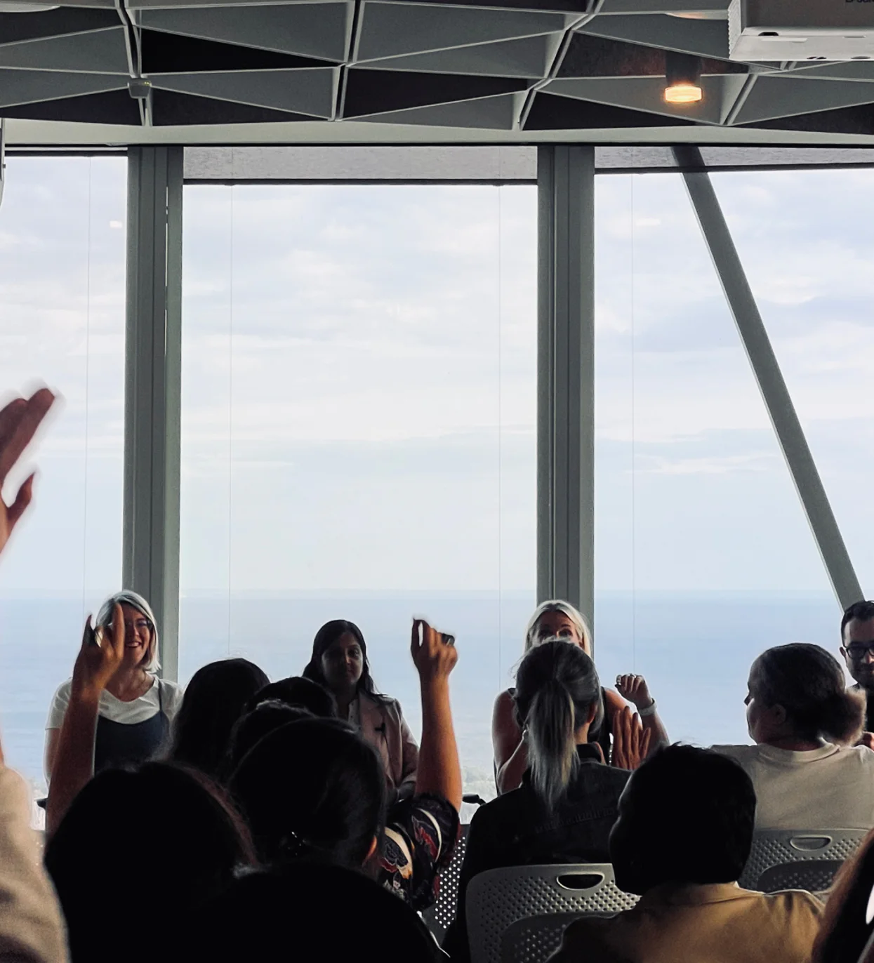 A group of people attending the a11yTO Meetup at Microsoft Toronto in a room with large windows overlooking a body of water. The attendees are seated, with some raising their hands, possibly to ask questions. The presenters or speakers are visible at the front, silhouetted against the bright view outside overlooking Lake Ontario.. 