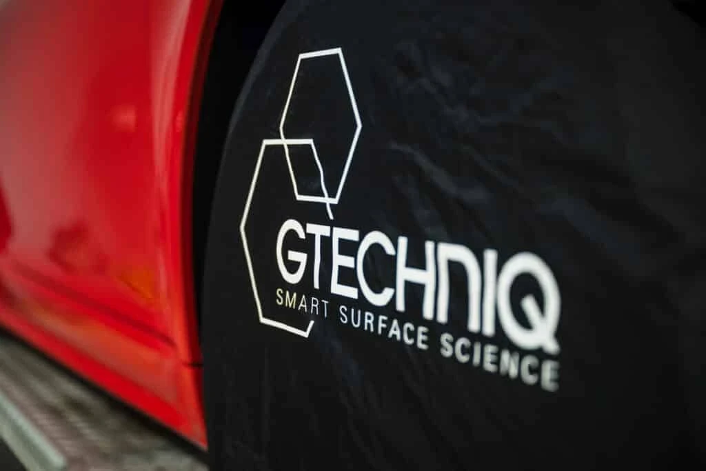 Close-up of a Gtechniq Smart Surface Science tire cover next to a glossy red car door, highlighting the brand’s focus on advanced automotive surface protection. The logo features hexagon outlines and bold white text.