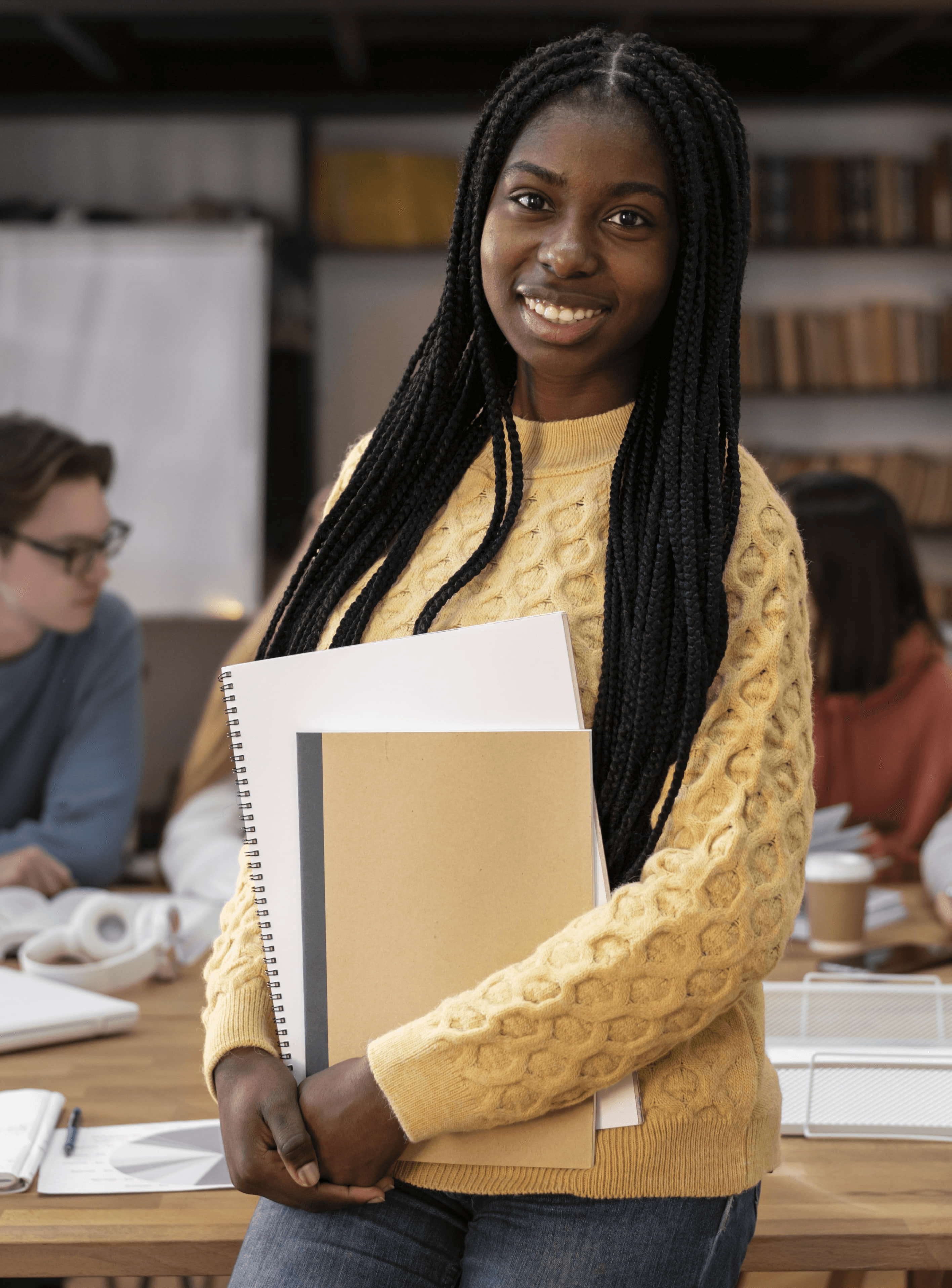 Smiling person in a yellow sweater holding documents, standing in a study space with others in the background.