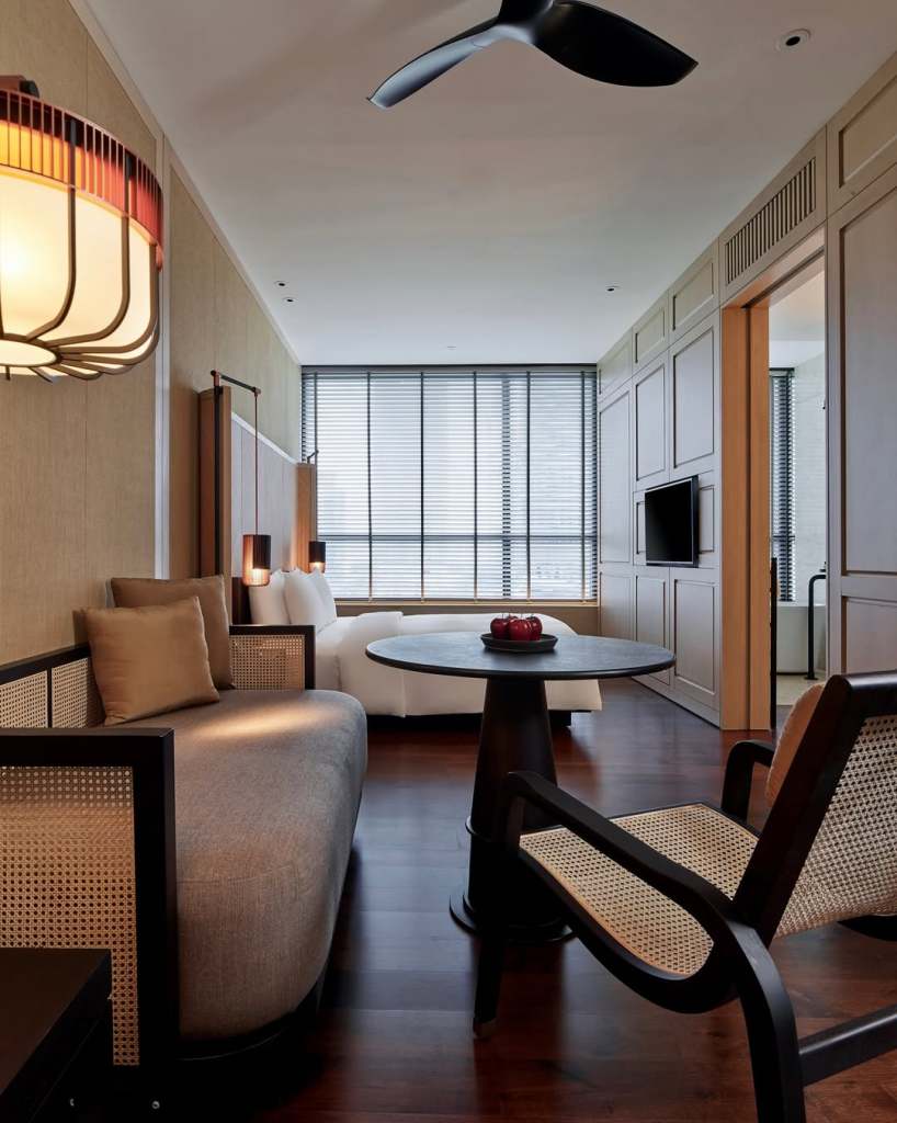 A modern hotel room featuring a cozy sitting area with a stylish beige sofa, a round table with apples, and a black ceiling fan, illuminated by soft lighting and large windows with blinds.
