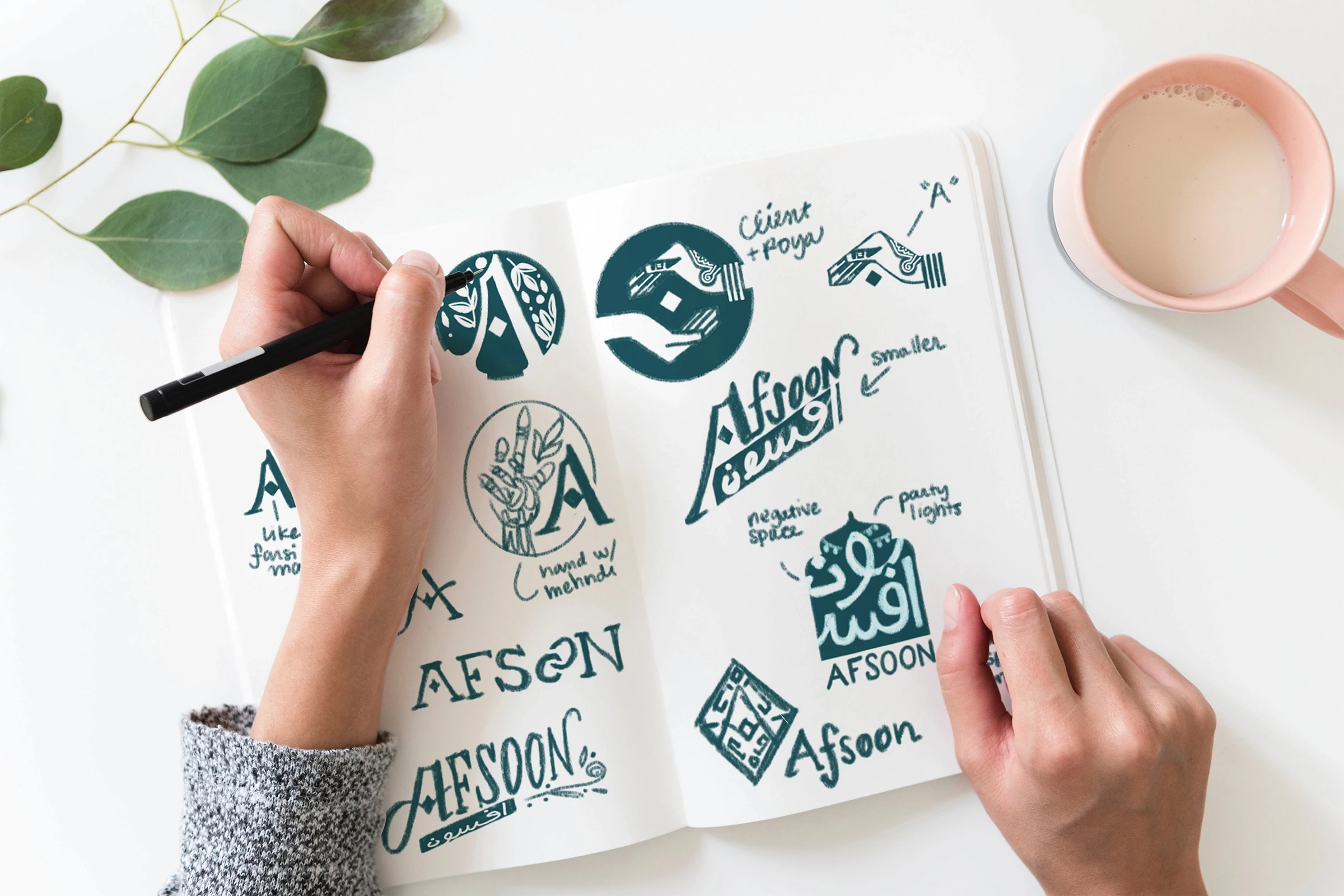 A pair of hands hold open a sketchbook. One hand holds a pencil, finishing a drawing of a logo idea alongside others across the pages.