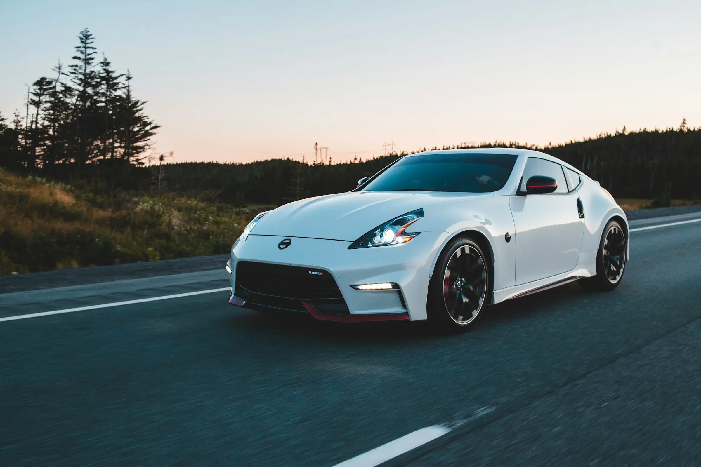 White sports coupe driving on an open highway at sunset with forest in the background