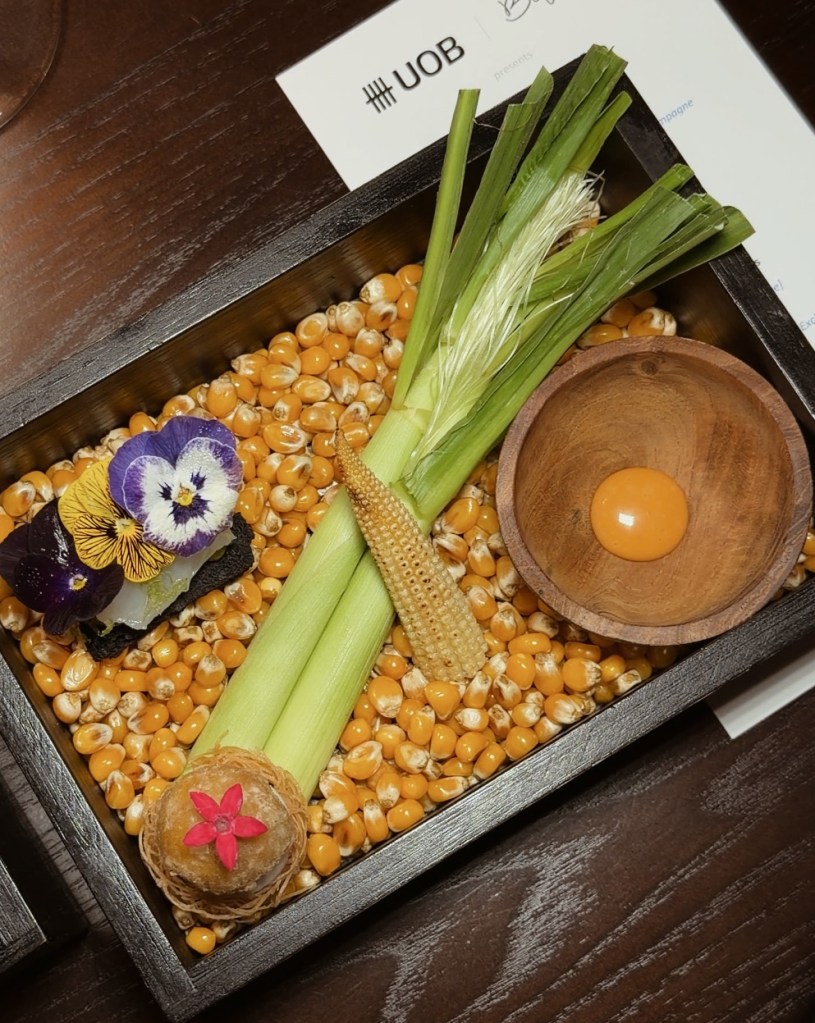 A wooden platter filled with yellow corn kernels, garnished with green vegetables, edible flowers, and a small bowl containing a yolk.