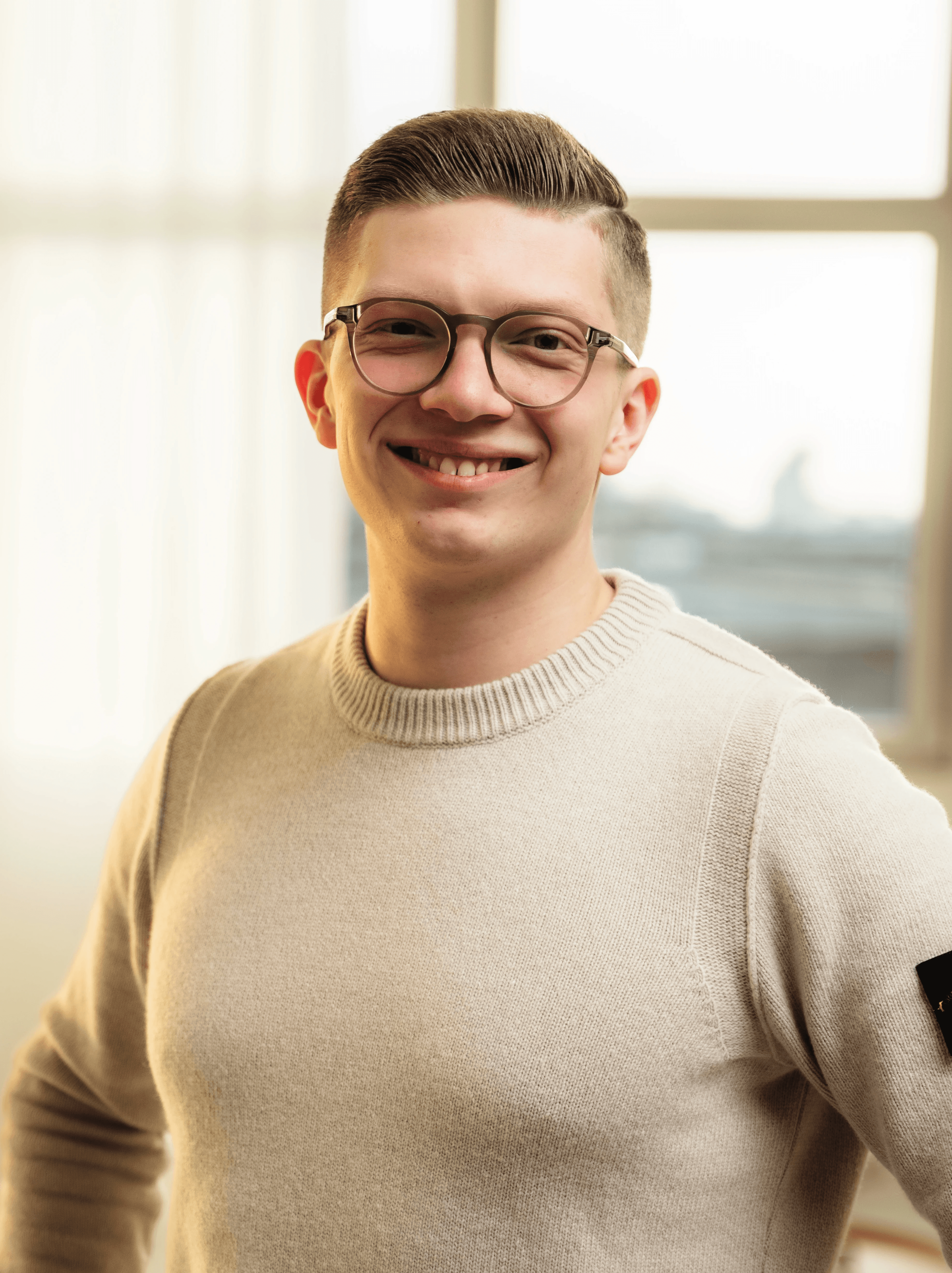 Close-up portrait of a person wearing a gray insulated hooded jacket with the hood up, showing their face framed by the hood against a light background.