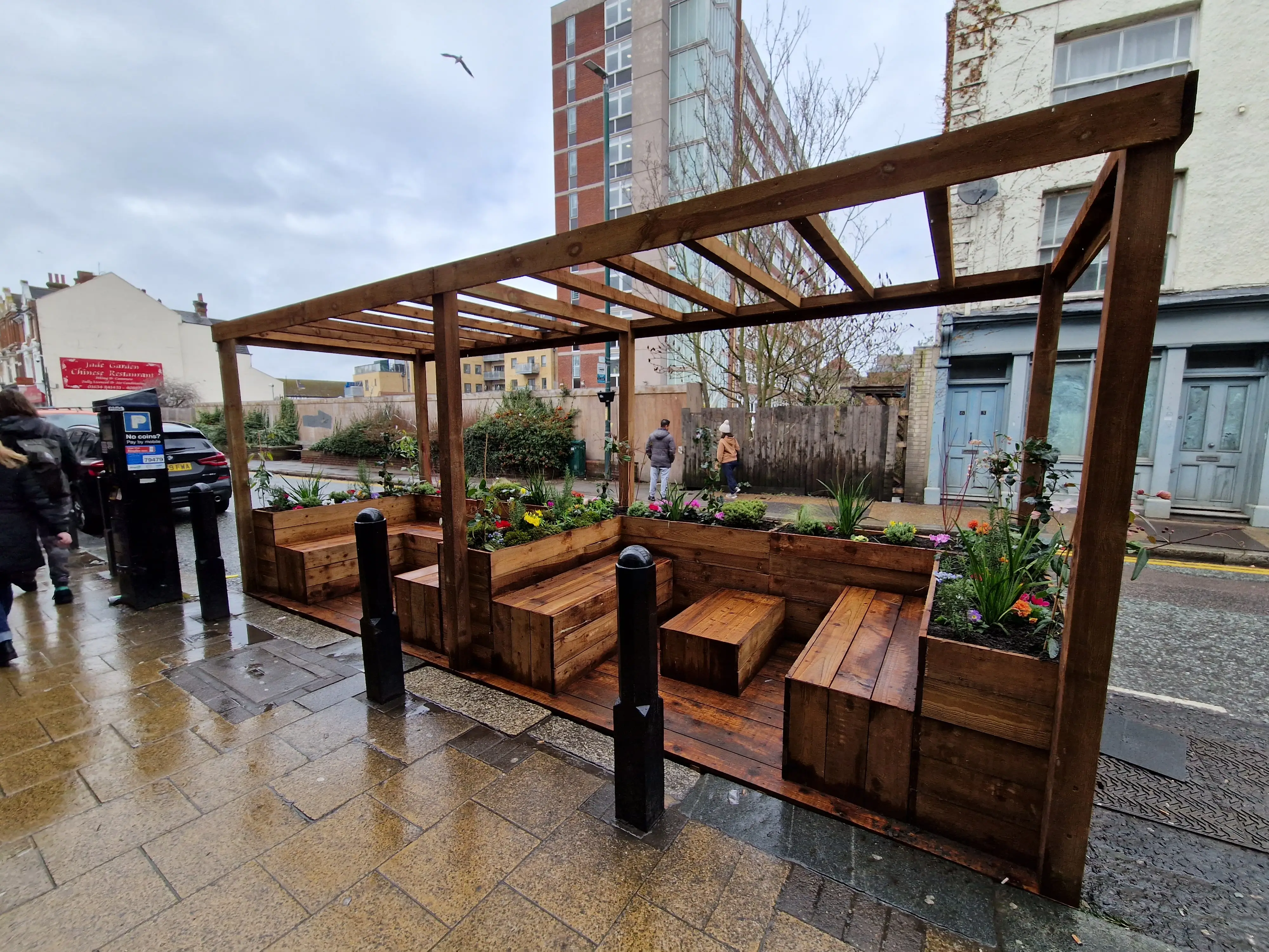 A wooden outdoor seating area with benches, under a pergola, on a rainy day in an urban setting.