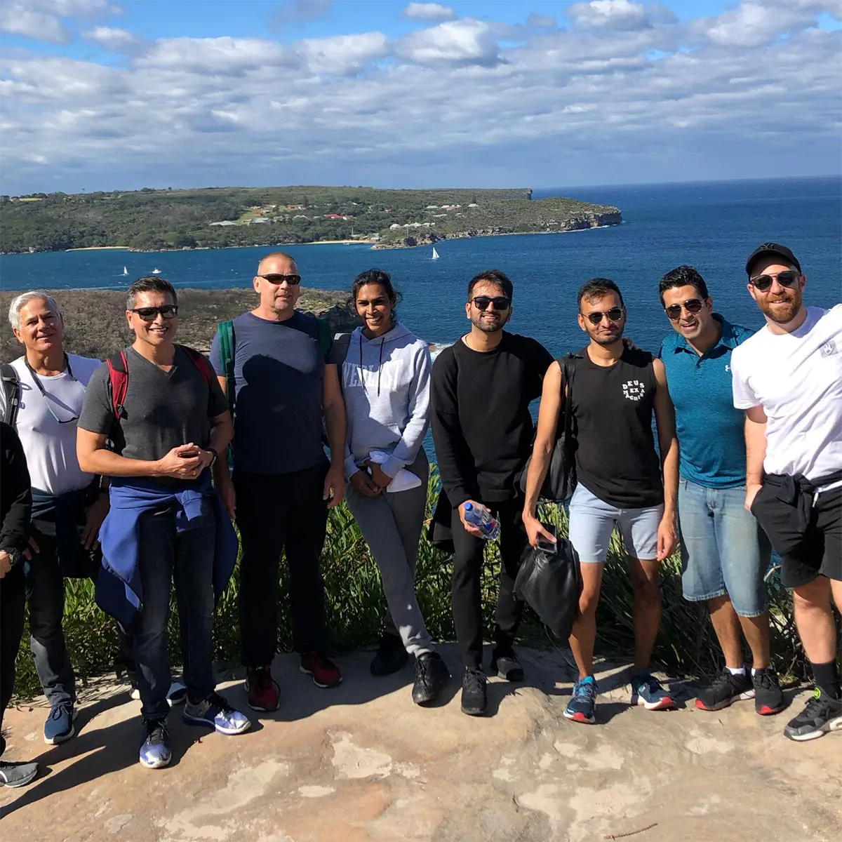 A group of eight people stands outdoors on a rocky spot with the sea and green coastline in the background under a partly cloudy sky. They are dressed casually and smiling at the camera.