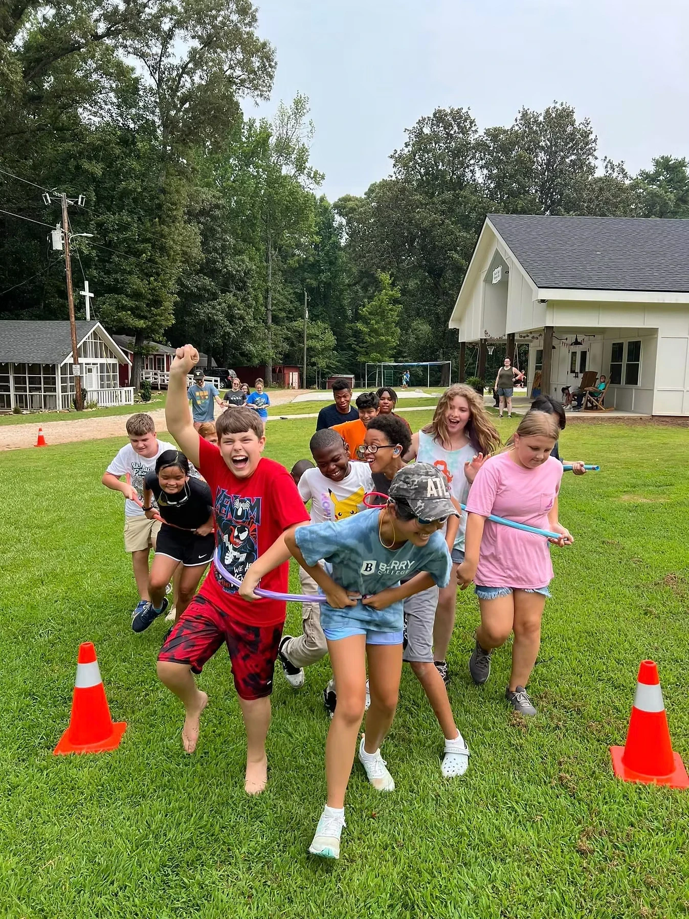 A group of kids playing a game outdoors on a grassy field. They are holding a long ribbon and running. Two orange cones are next to them.