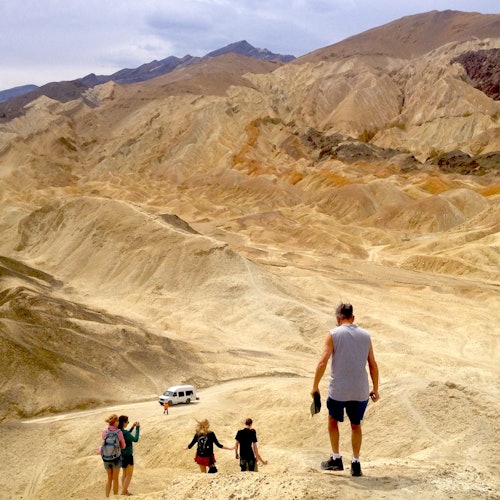 A group of people hiking down a sandy, hilly terrain with a white van parked in the background. Mountains are visible under an overcast sky.