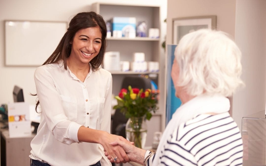 A young woman shakes hands with an older woman in an inviting office setting. Flowers are seen in the background.