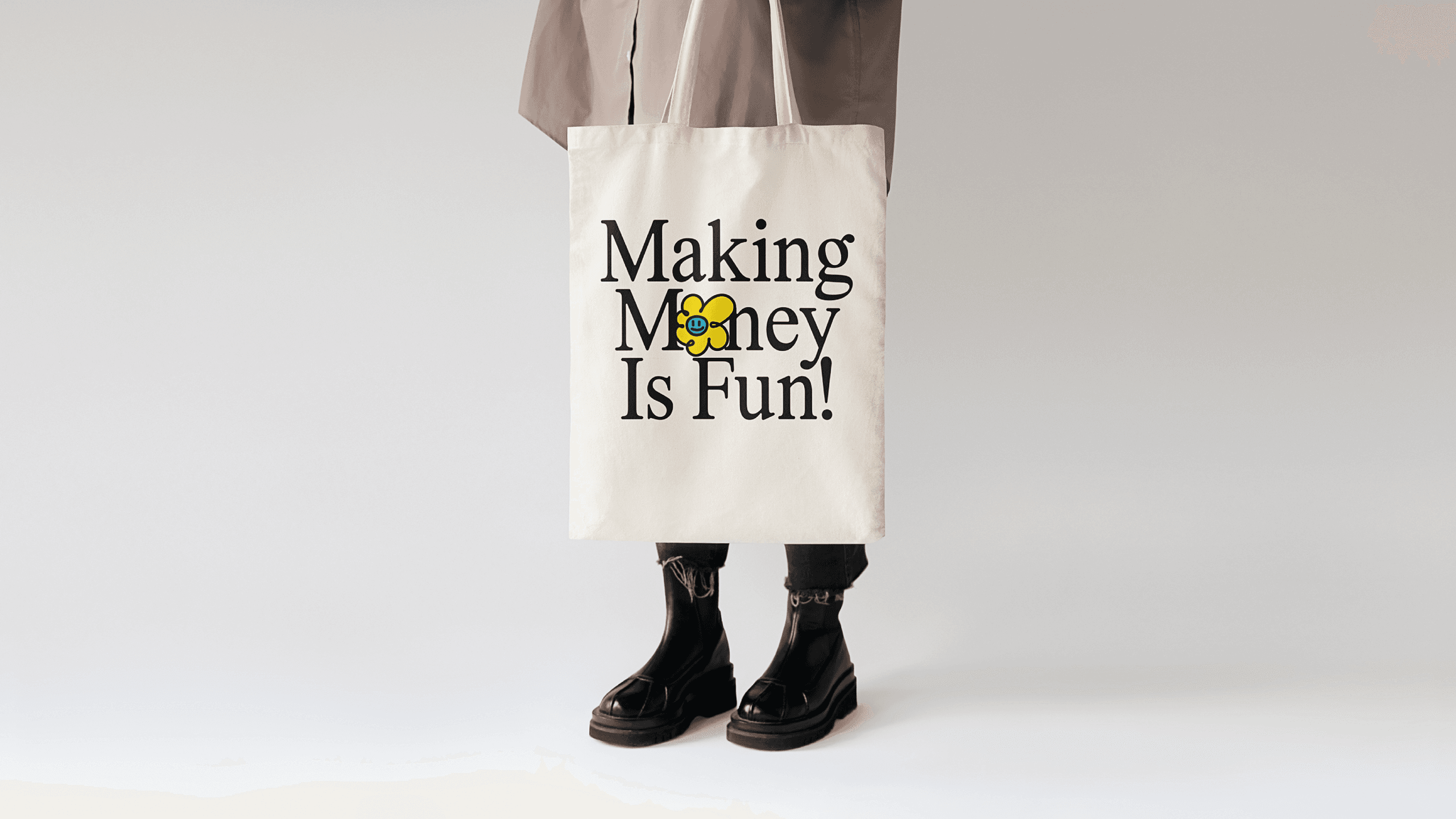 A person stands against a plain light gray background, wearing a gray coat, black pants, and black boots. They are holding a white tote bag with the text "Making Money Is Fun!" printed in black letters. A small yellow flower with a yellow center is incorporated into the text.