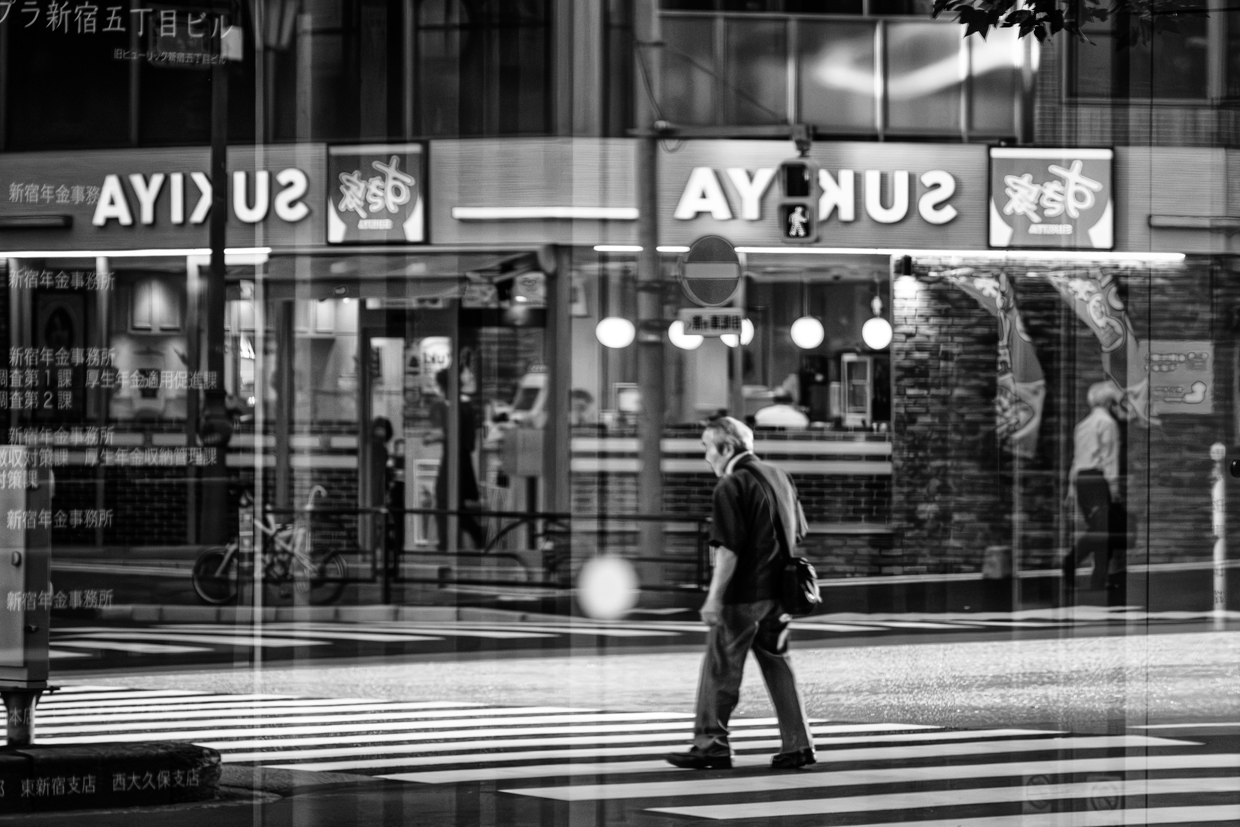 Black and white photograph of commuters seen through a train window with reflections of another person on the station platform overlaying the scene.