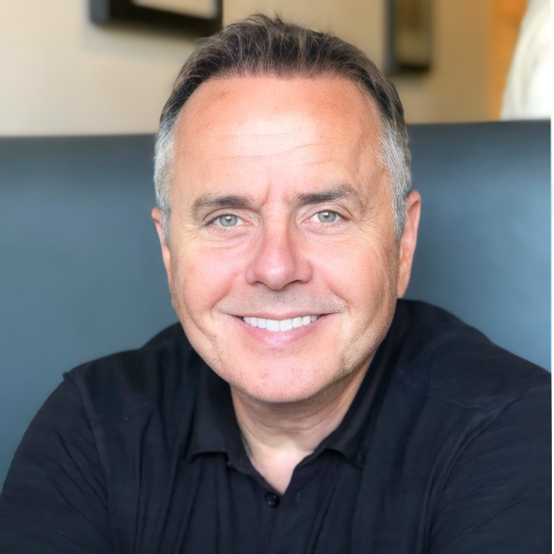 A smiling person with curly hair, wearing a leather jacket, stands in a cozy indoor setting.