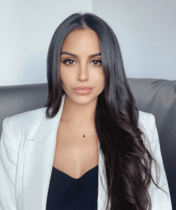 Smiling woman in beige yoga outfit sitting in natural light with green plants in the background.